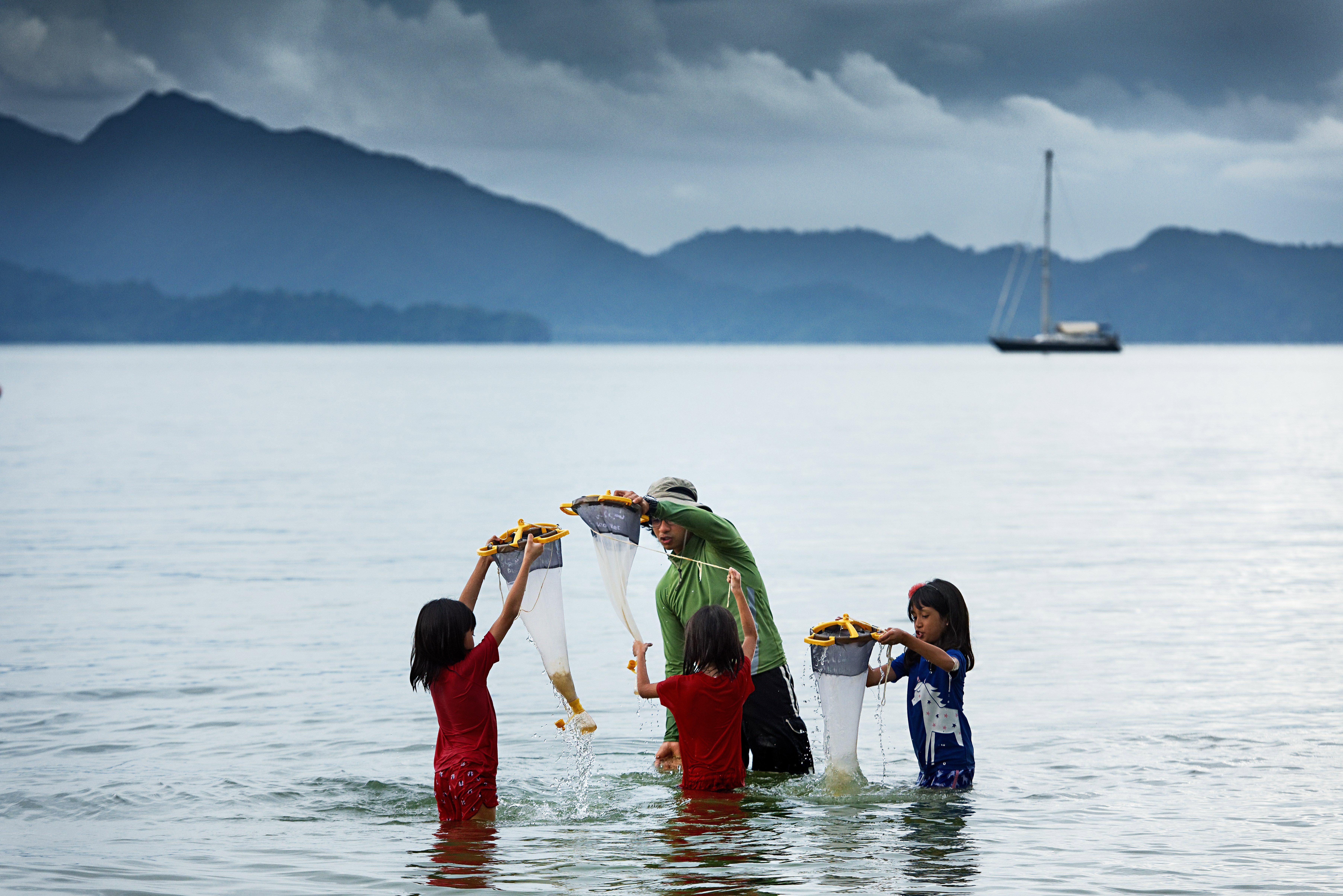 An adult and three children catching seafood in the calm waters of Langkawi with mountains and a boat drifting in the distance
