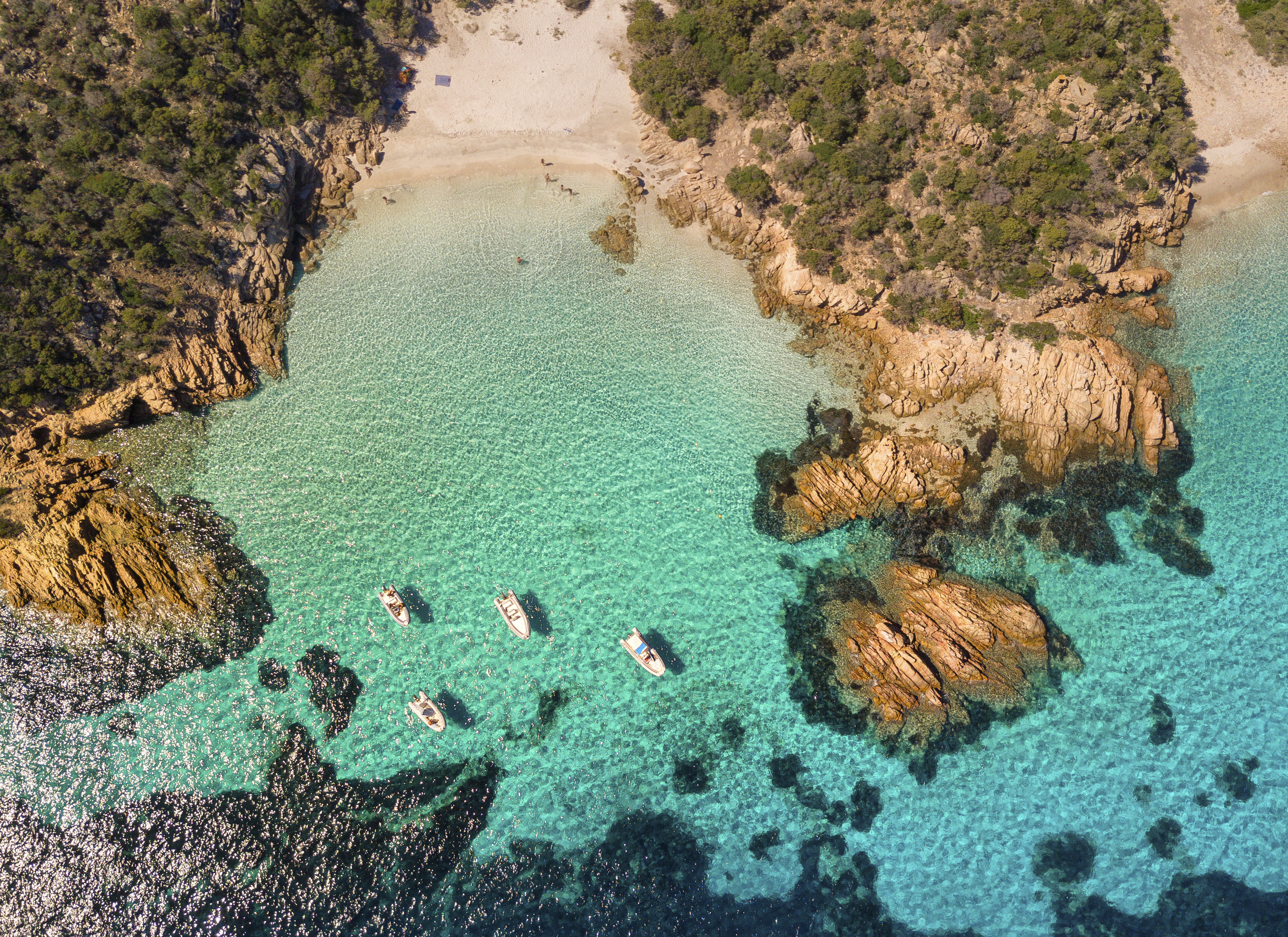 Aerial view of a clear turquoise sea with boats near a rocky coastline and sandy beach.