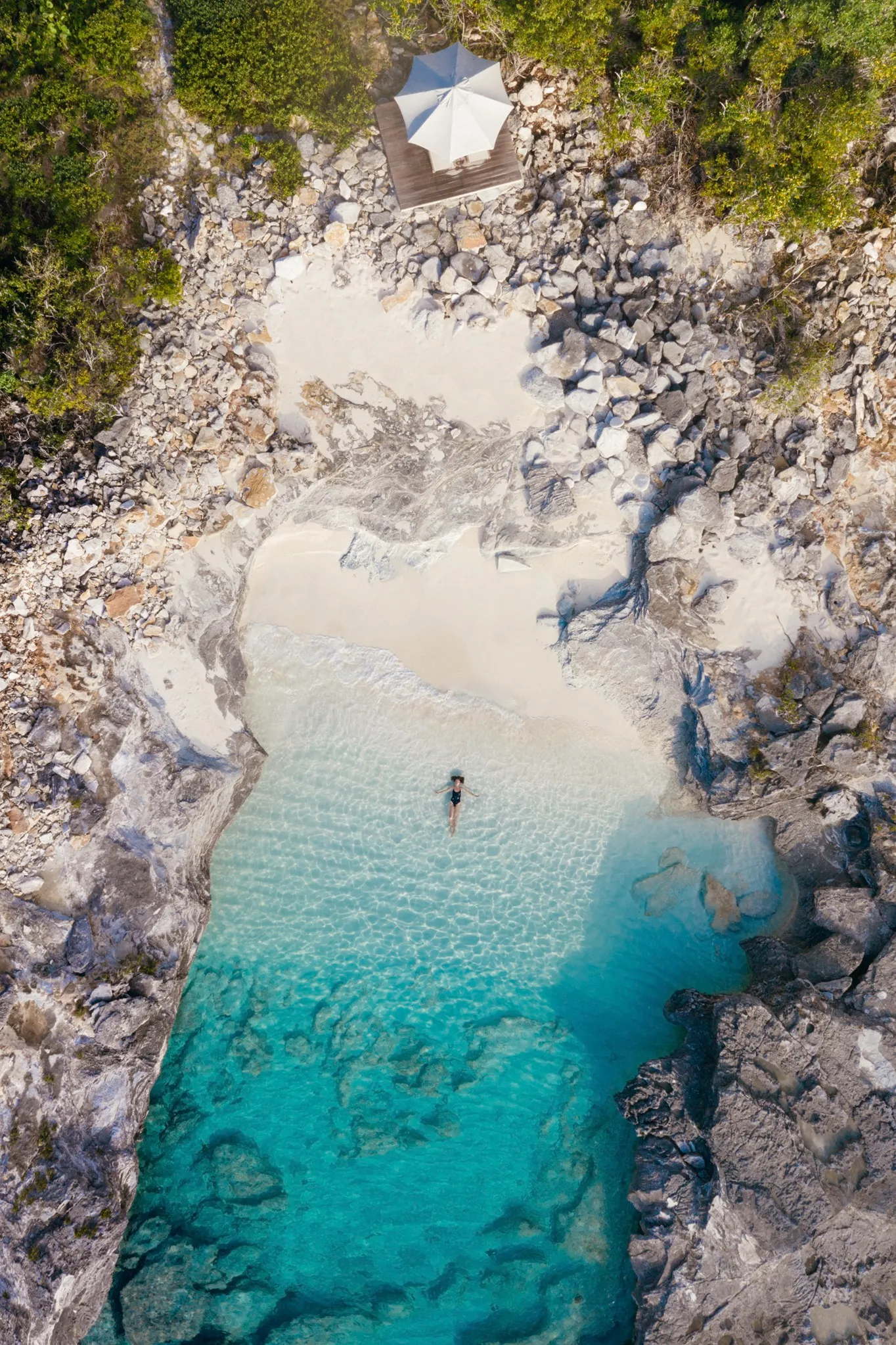 A lies in the shallows in a clear turquoise cove surrounded by rocky cliffs and greenery, with a white canopy on the shore.