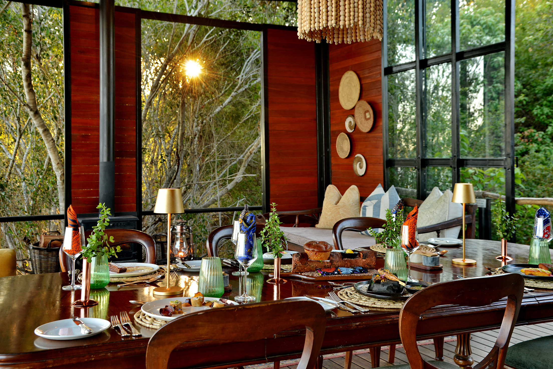 A sunlit dining table elegantly set with dishes, plants, and decor, surrounded by trees and a cozy seating area at Mukwa River Lodge
