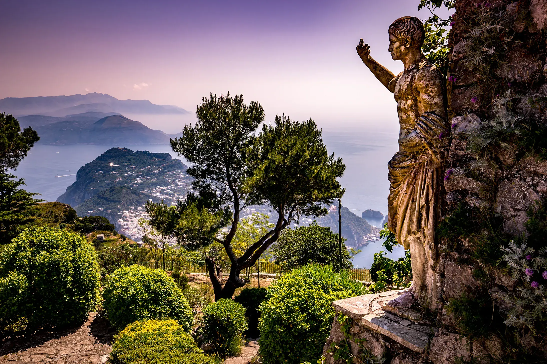 Scenic view of Capri coastline from a garden with a historic statue and lush greenery.