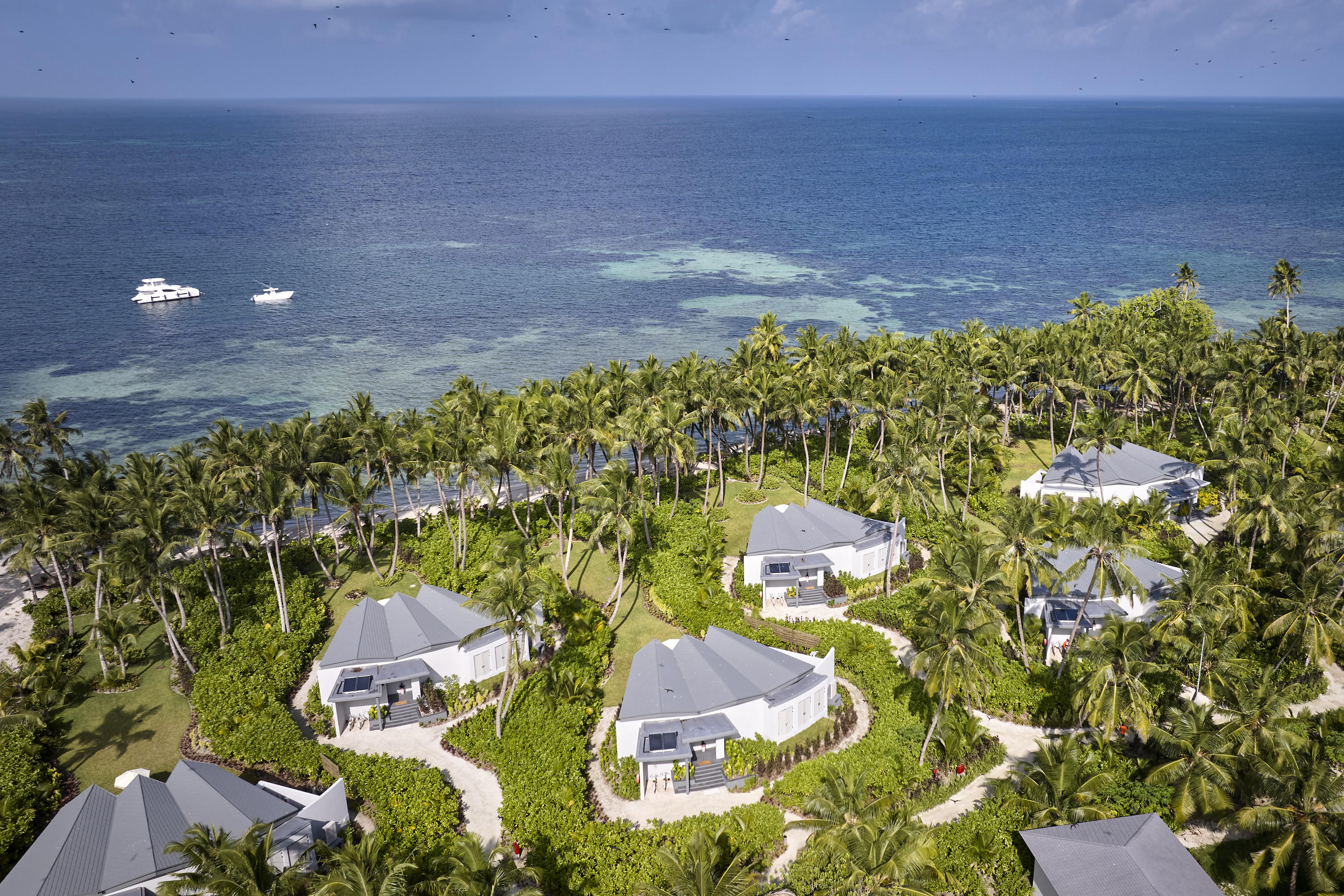 Birds eye view of pool villas and gardens looking out to the ocean where a white yacht is anchored 