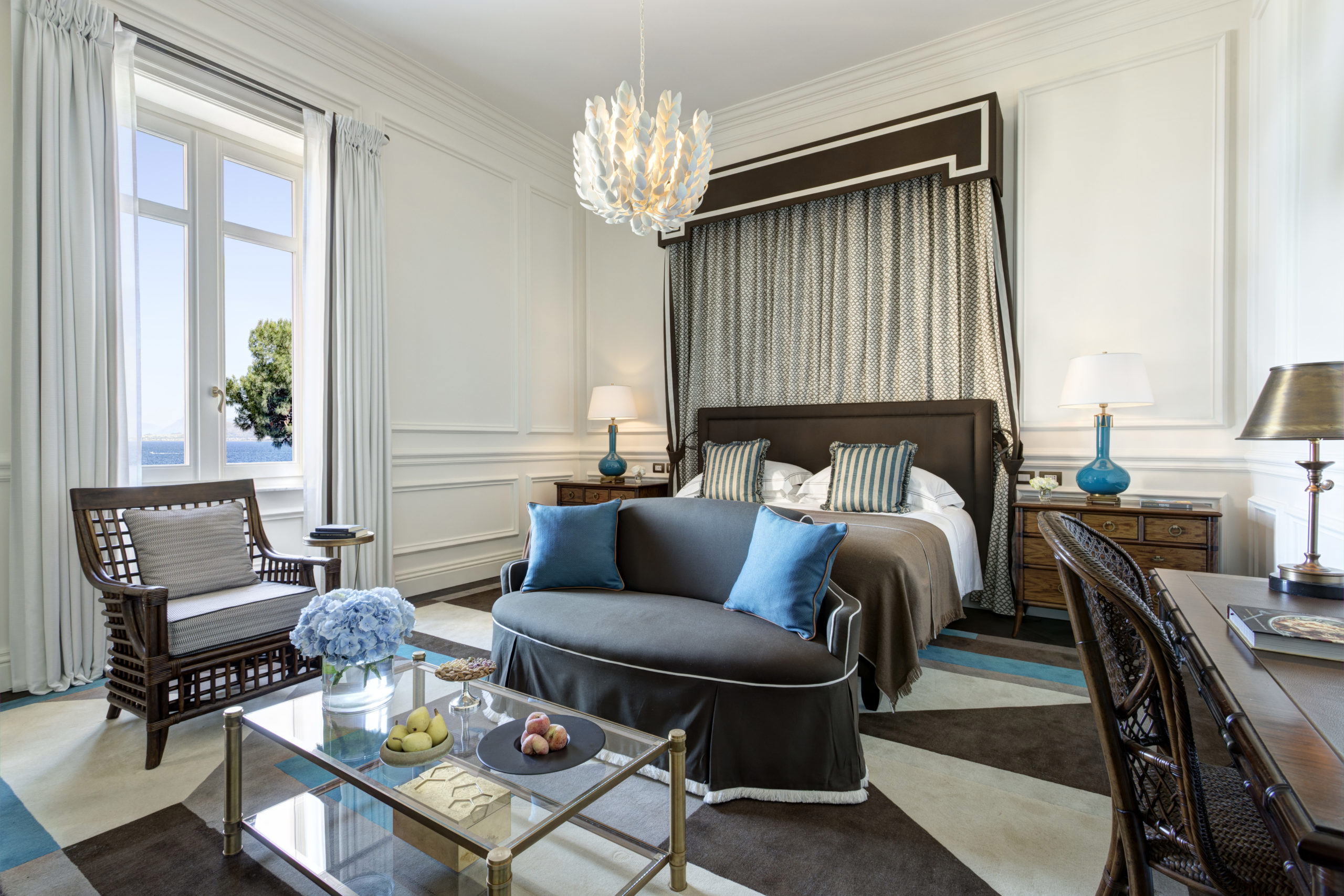 An elegantly decorated bedroom with a large bed, two bedside lamps, a chandelier, and a seating area with chairs and a glass coffee table.