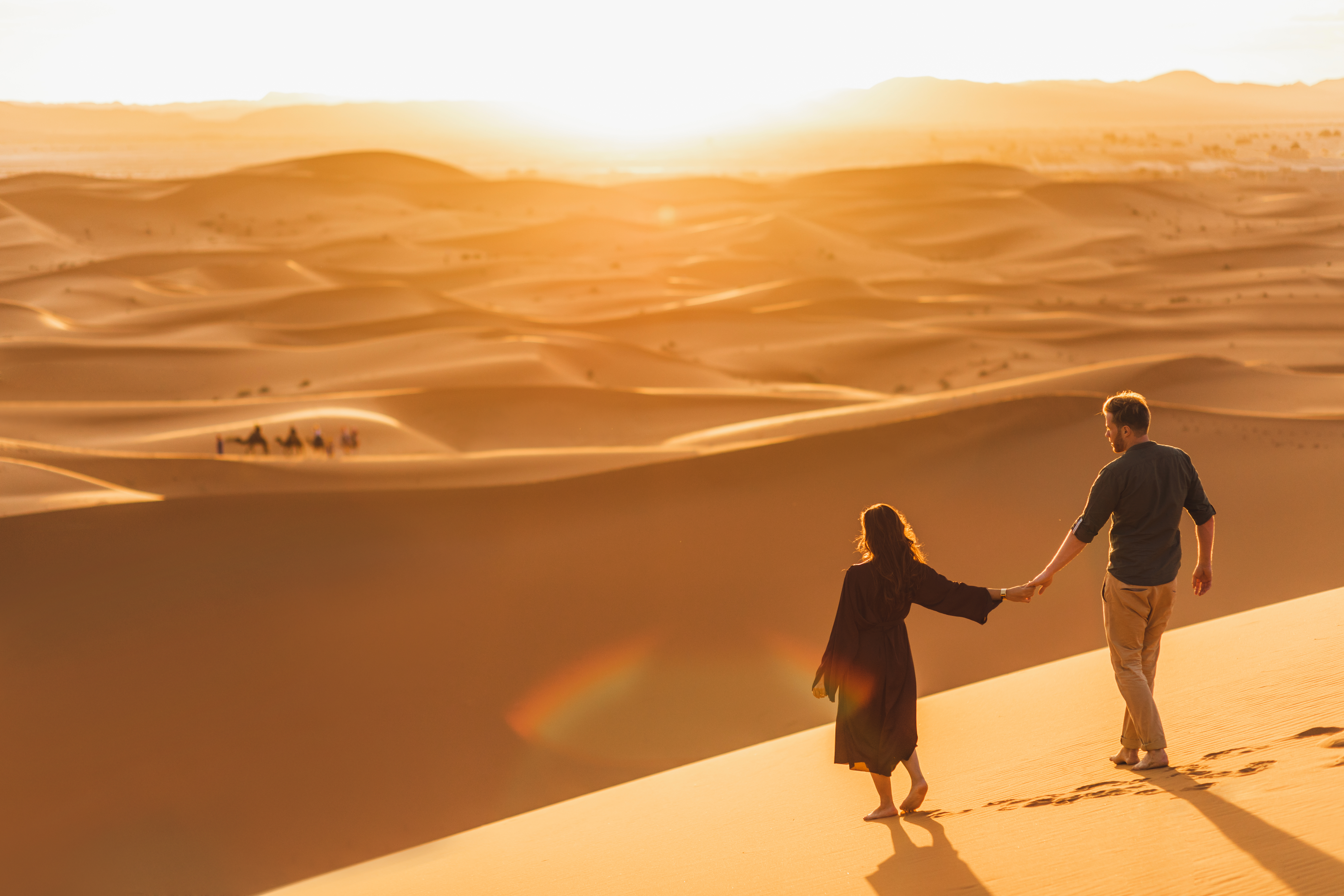 A couple holding hands walks on sand dunes at sunset in a vast desert landscape, with camels seen in the background.