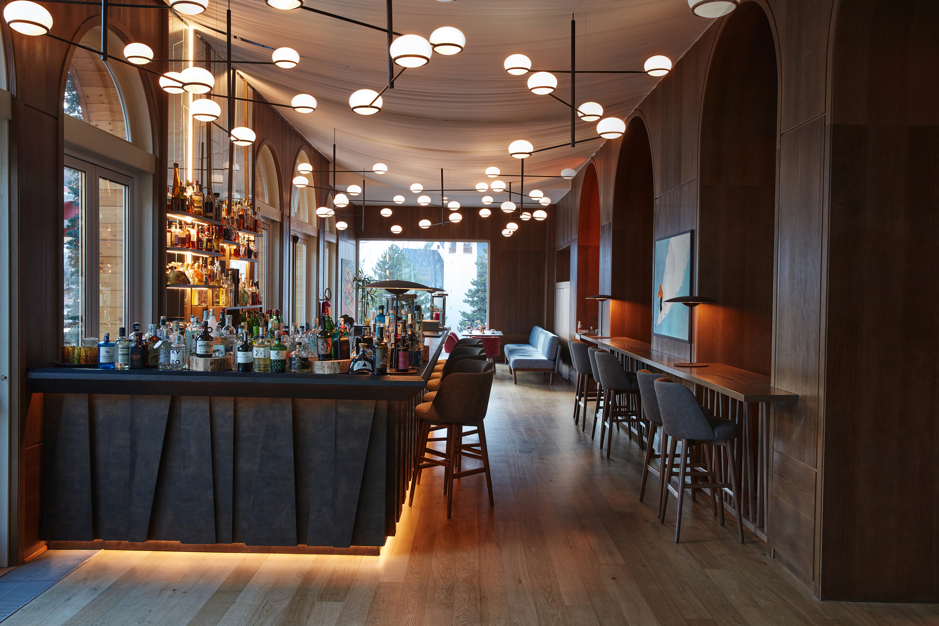The lounge bar of Grand Hotel Savoia featuring a sleek modern bar lined with bottles and stools with more seating opposite and statement lighting