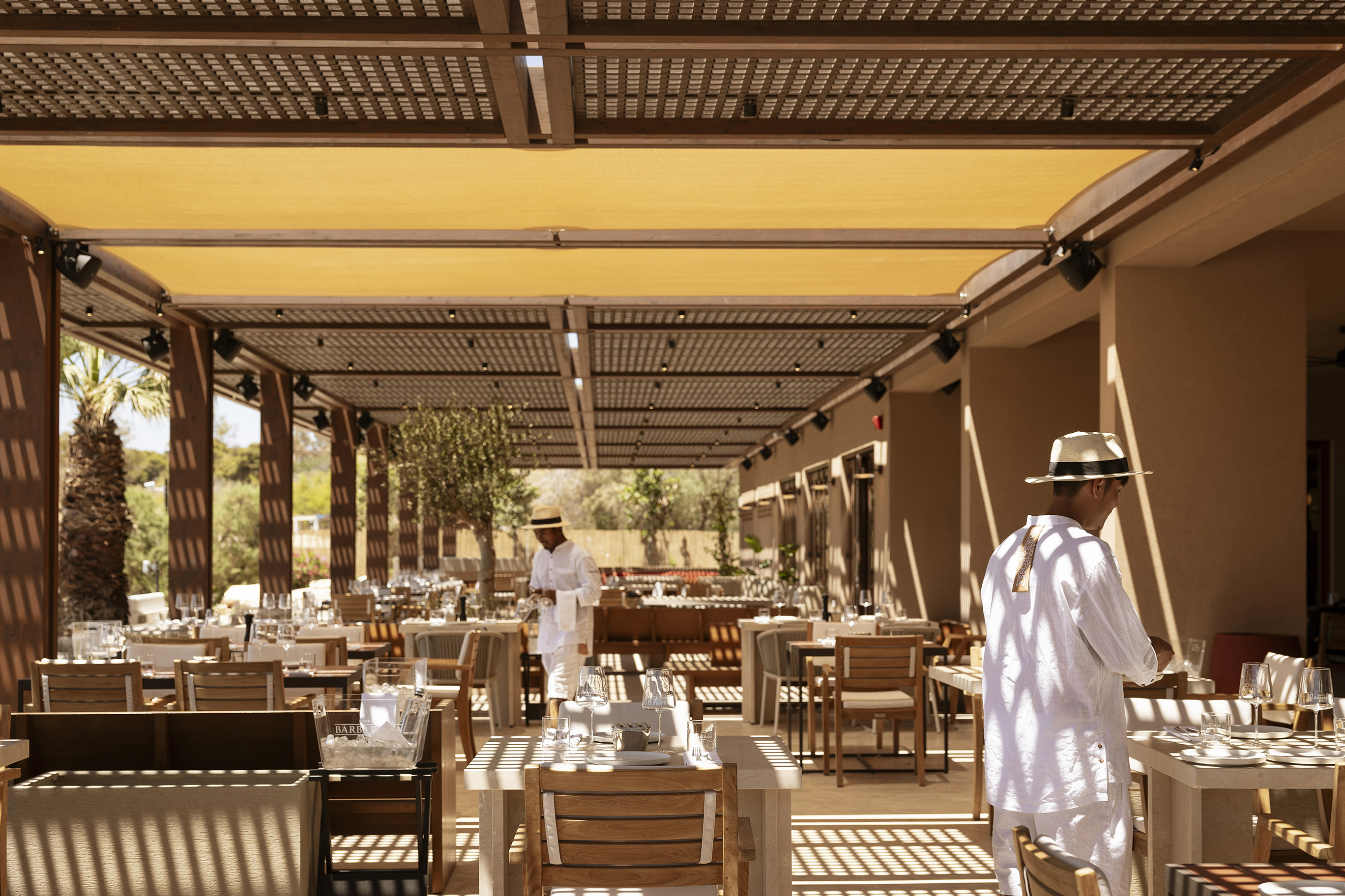Wooden dining tables set below a wooden cabana with two men dressed in white with sun hats