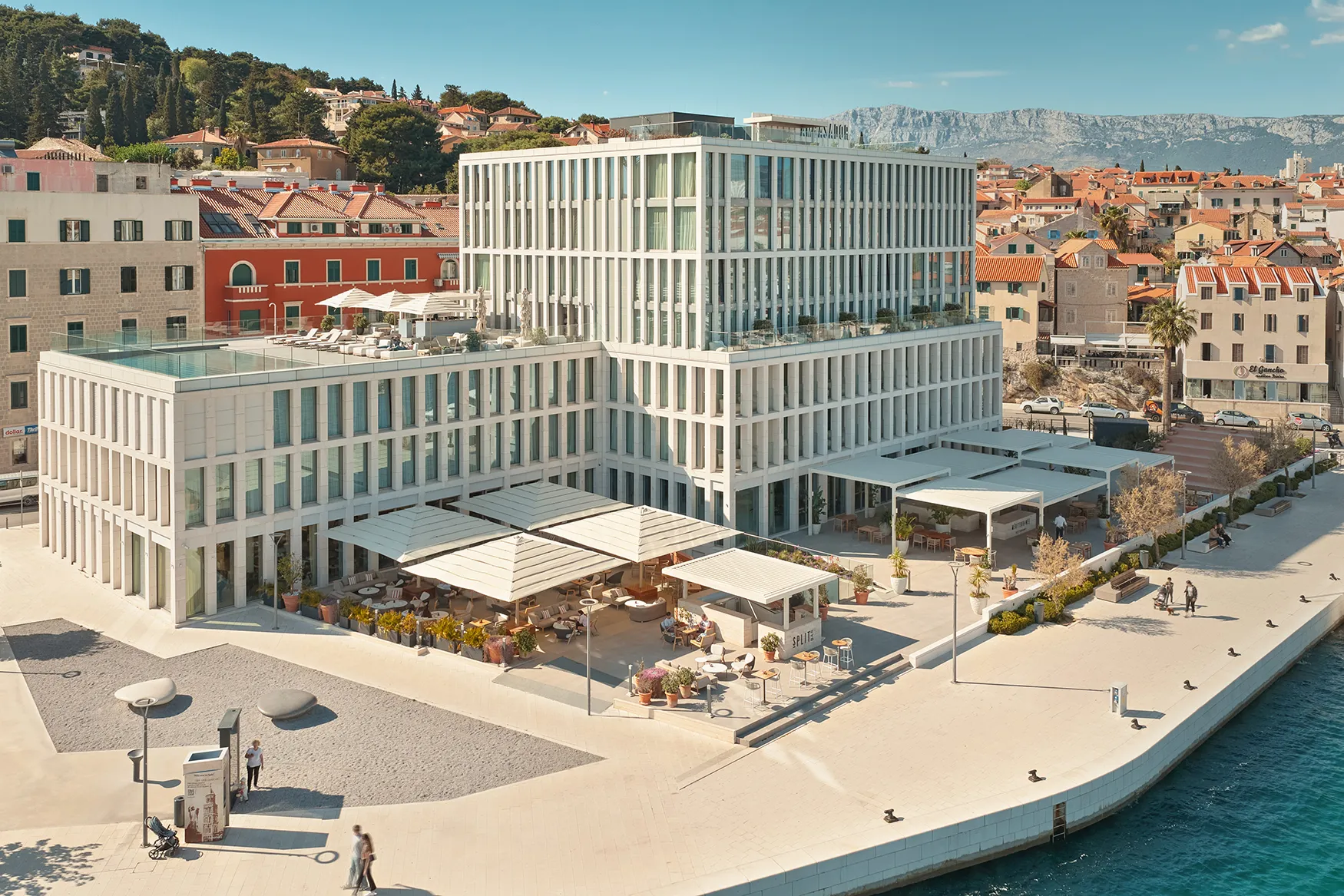 Modern waterfront hotel with glass façades and outdoor terraces, set beside a promenade and historic coastal buildings.