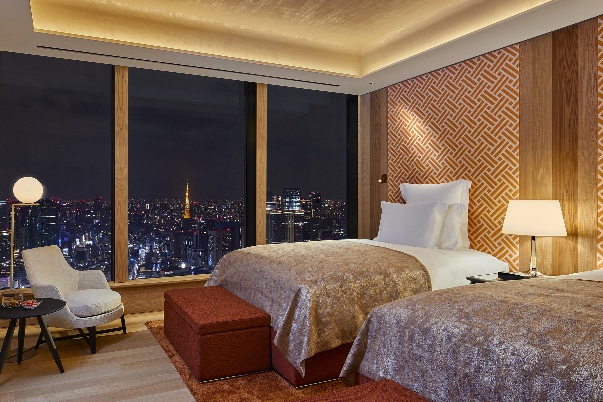 A twin room at the Bulgari Tokyo lit up at night featuirng two beds with pink throws and a comfy seat with a view of the city
