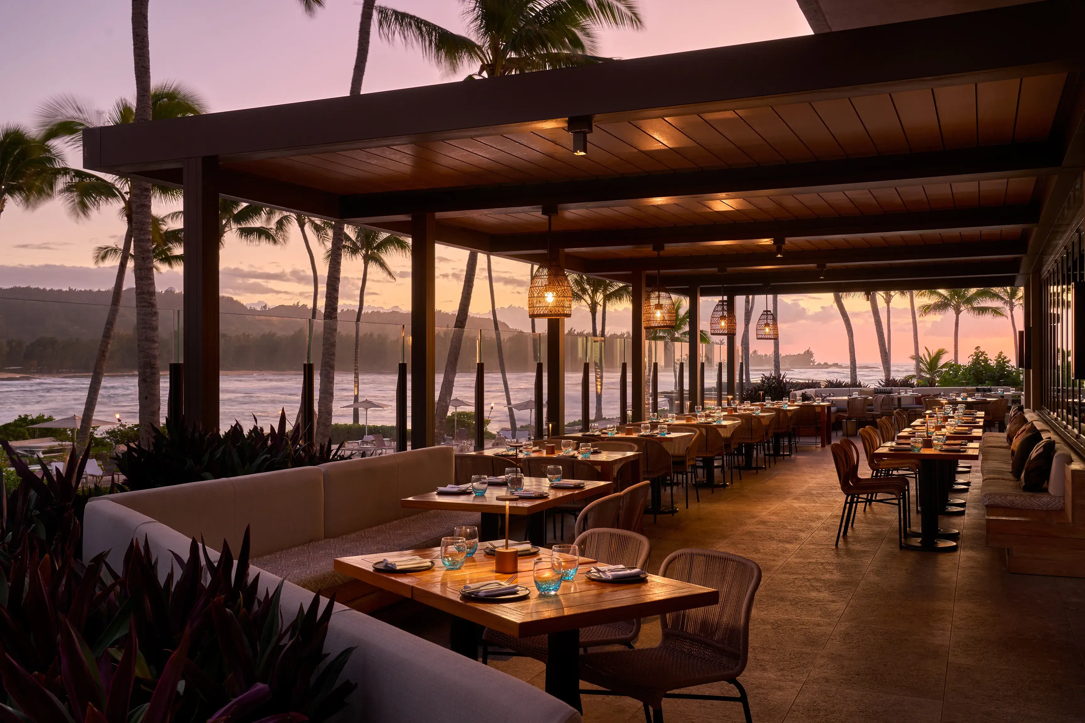 The indoor/outdoor terrace of Alaia Restaurant at Ritz-Carlton Turtle Bay at sunset, featuring tables set up for dinner beneath cosy lighting backed by palm trees and the ocean