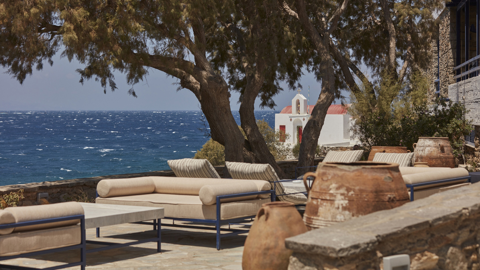 A comfy outdoor seating area shaded from the sun by a tree with decorative pots and a view of a small church through the branches