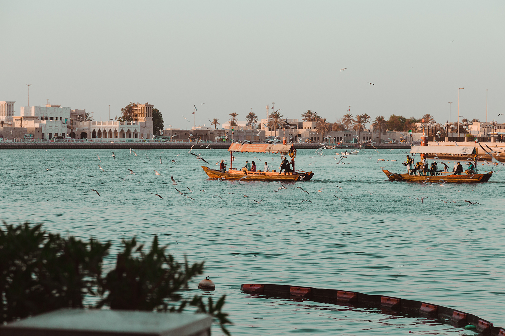 Traditional wooden boats on a calm river with flying birds and buildings in the background during sunset.
