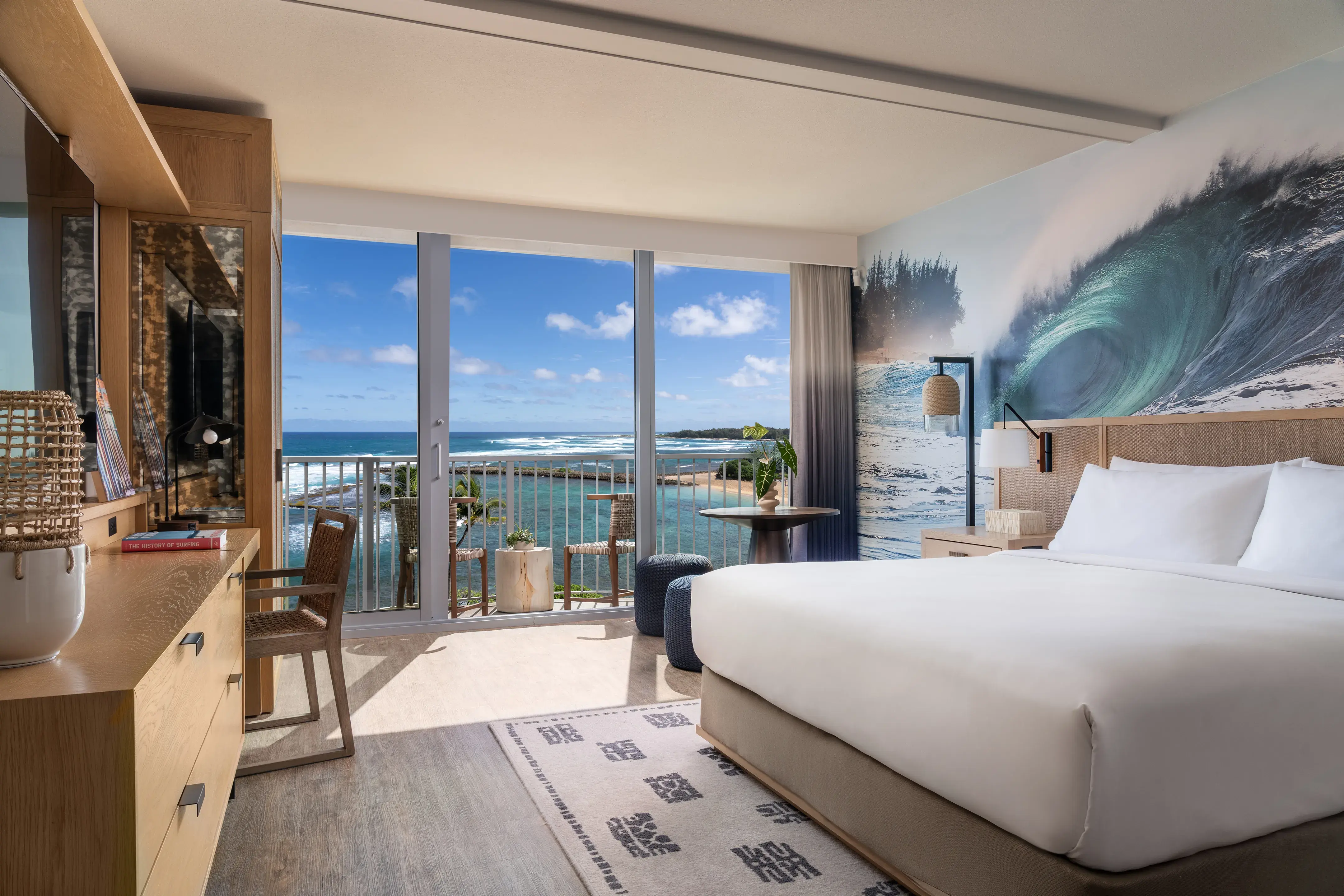 A Premium Oceanfront King Juliet at Ritz-Carlton Turtle Bay featuring a comfy bed in front of a wall decorated with a wave mural, with a writing area in front and large balcony doors looking out over the ocean