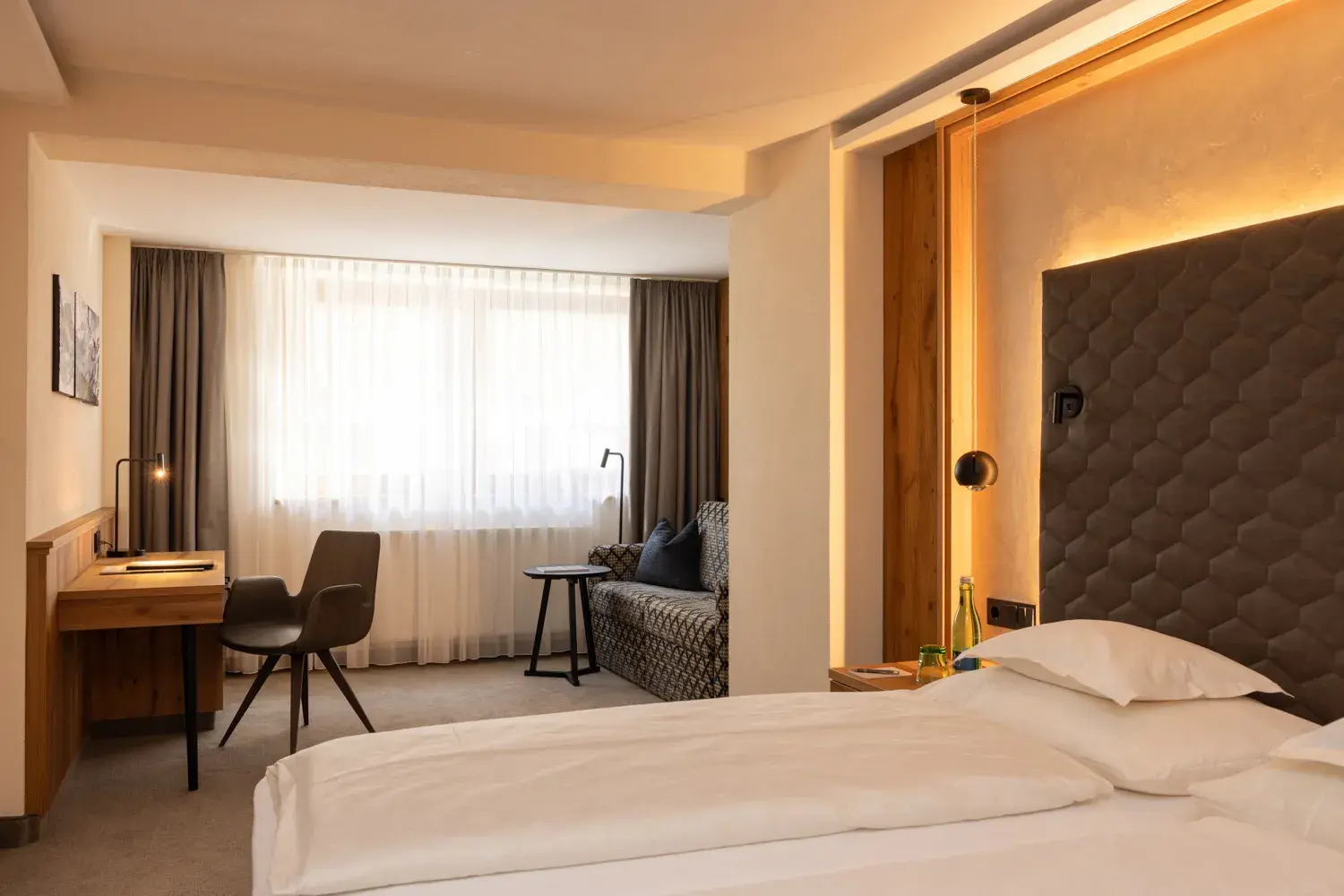 A spacious hotel room with two neatly made beds, a flat-screen TV, a wooden desk, and a sitting area with a sofa and large windows at Gourmet and Wine Hotel Austria