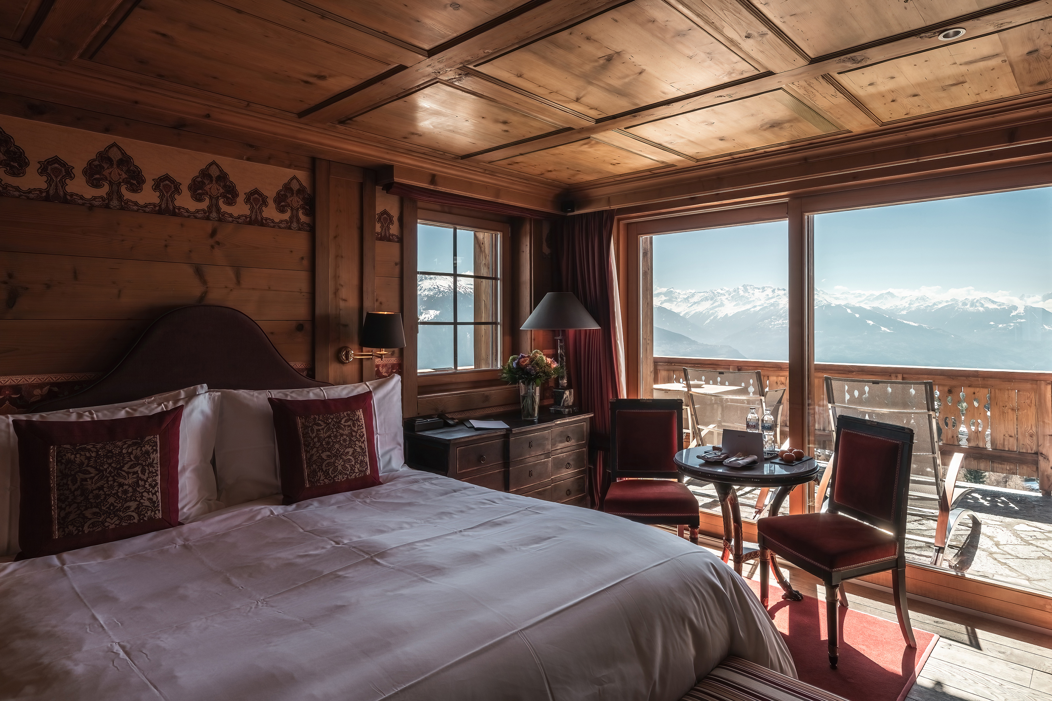 A cosy suite at LeCrans Hotel featuring a bed with ornate cushions and a small table and chair set, with large windows looking out onto a balcony with mountain views