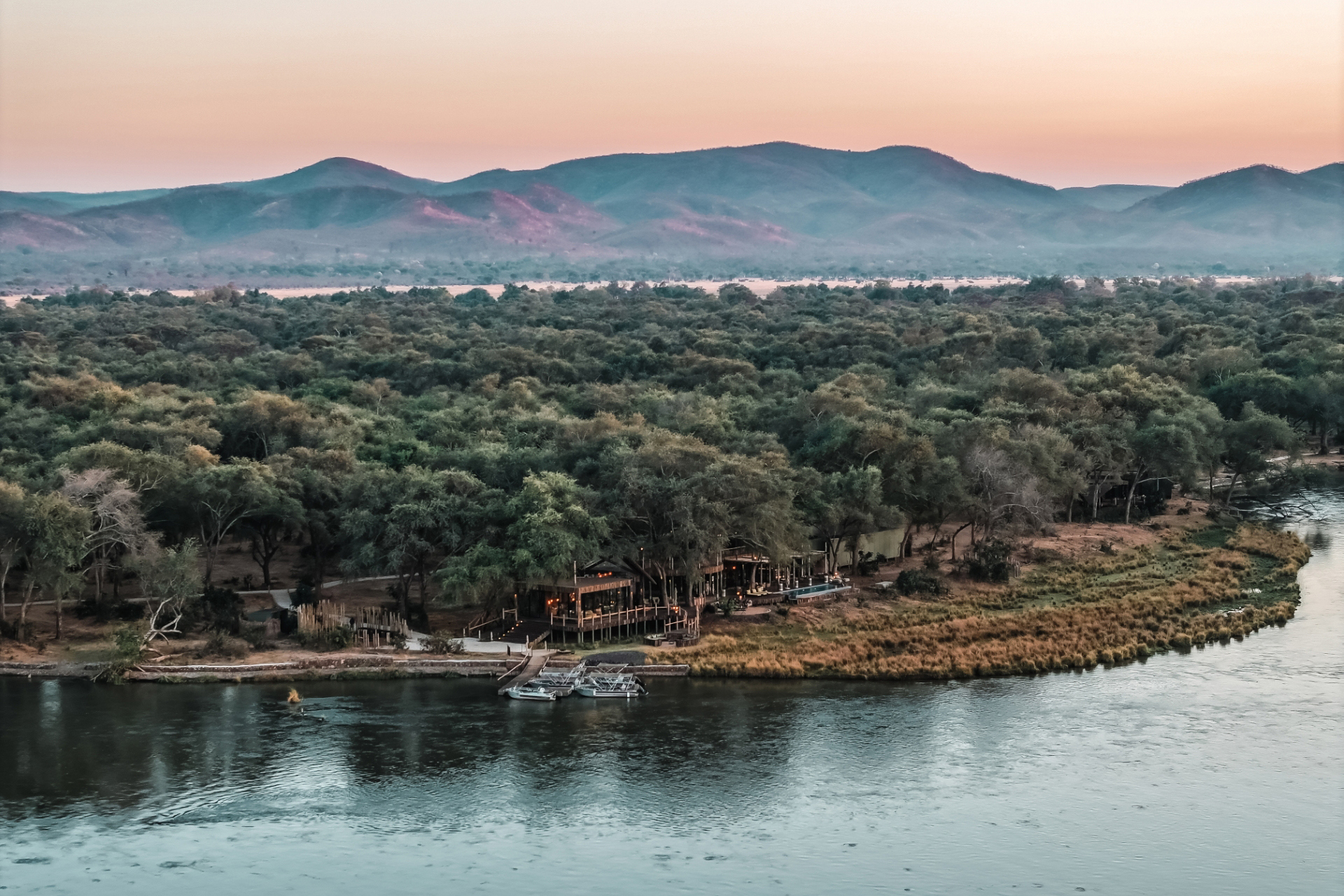 The vast landscape surrounding Lolebezi with a glimpse of the lodge by the river between the trees under a colourful sky at sunset