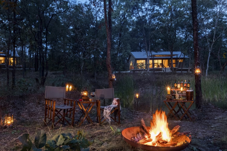 Two chairs and a drinks cabinet set around a fire pit in the jungle with a lodge lit up at night in the background
