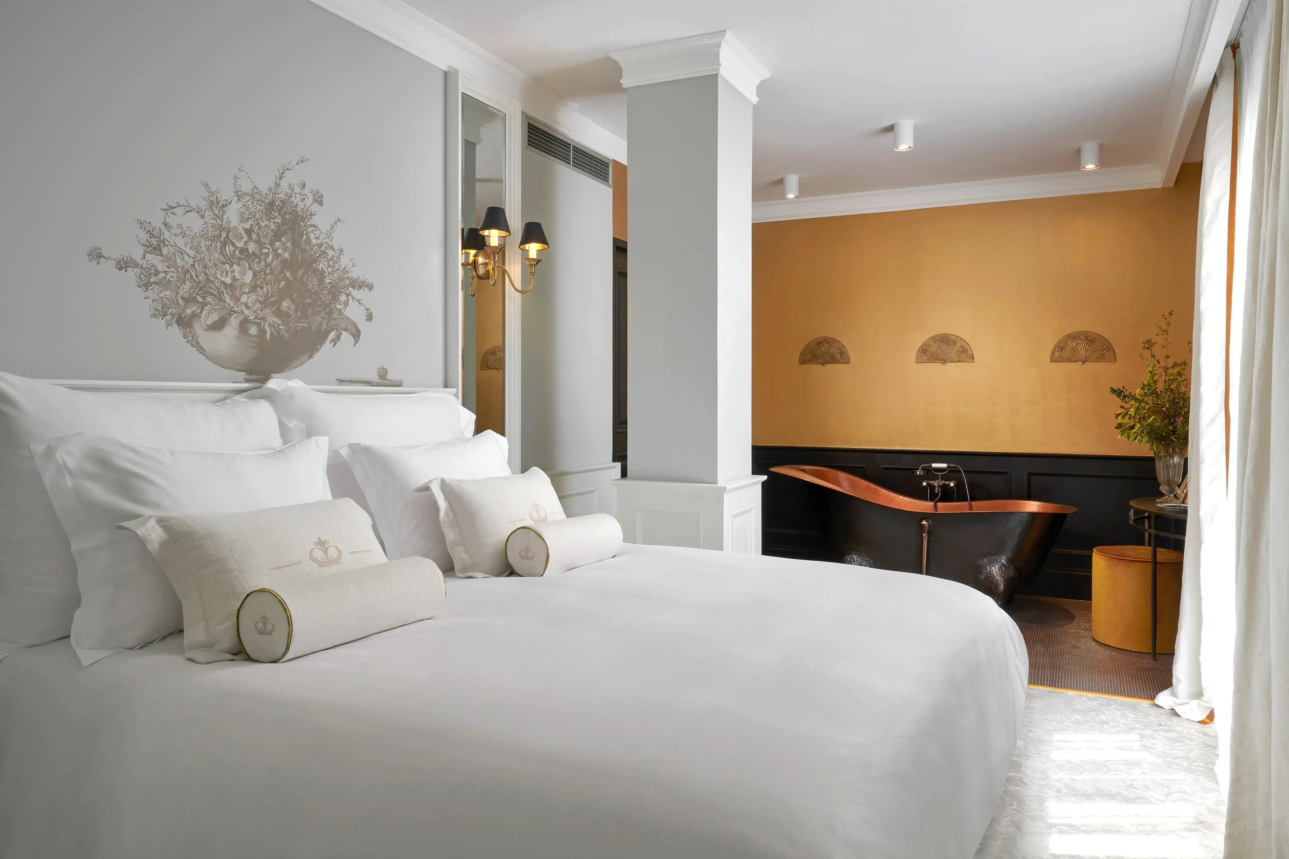 A Balcony Suite at Palace Elisabeth in Croatia featuring an all white bed area with plush bed and cushions with a gold bath area behind featuring a black and copper bathtub and gold walls
