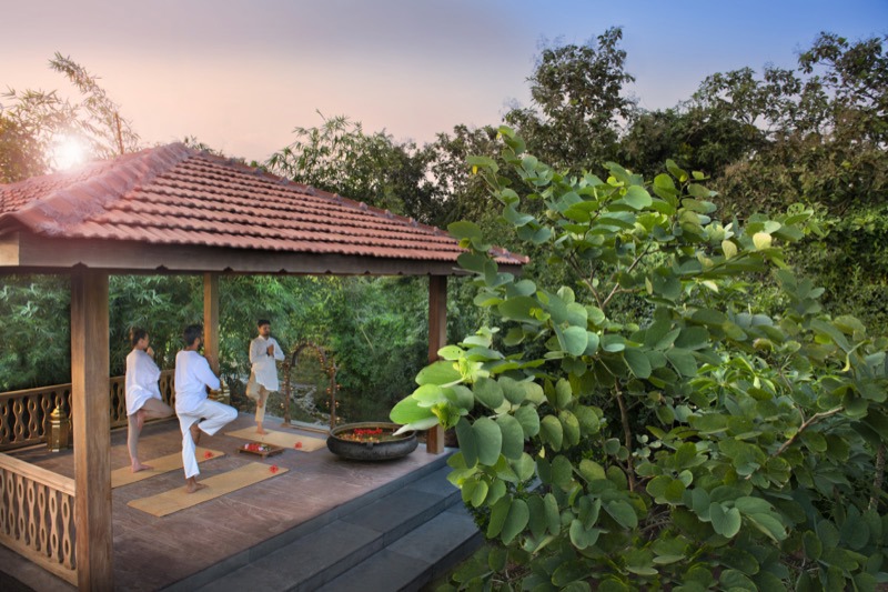 Three people in white practicing yoga in the red-roofed pavilion beside green foliage