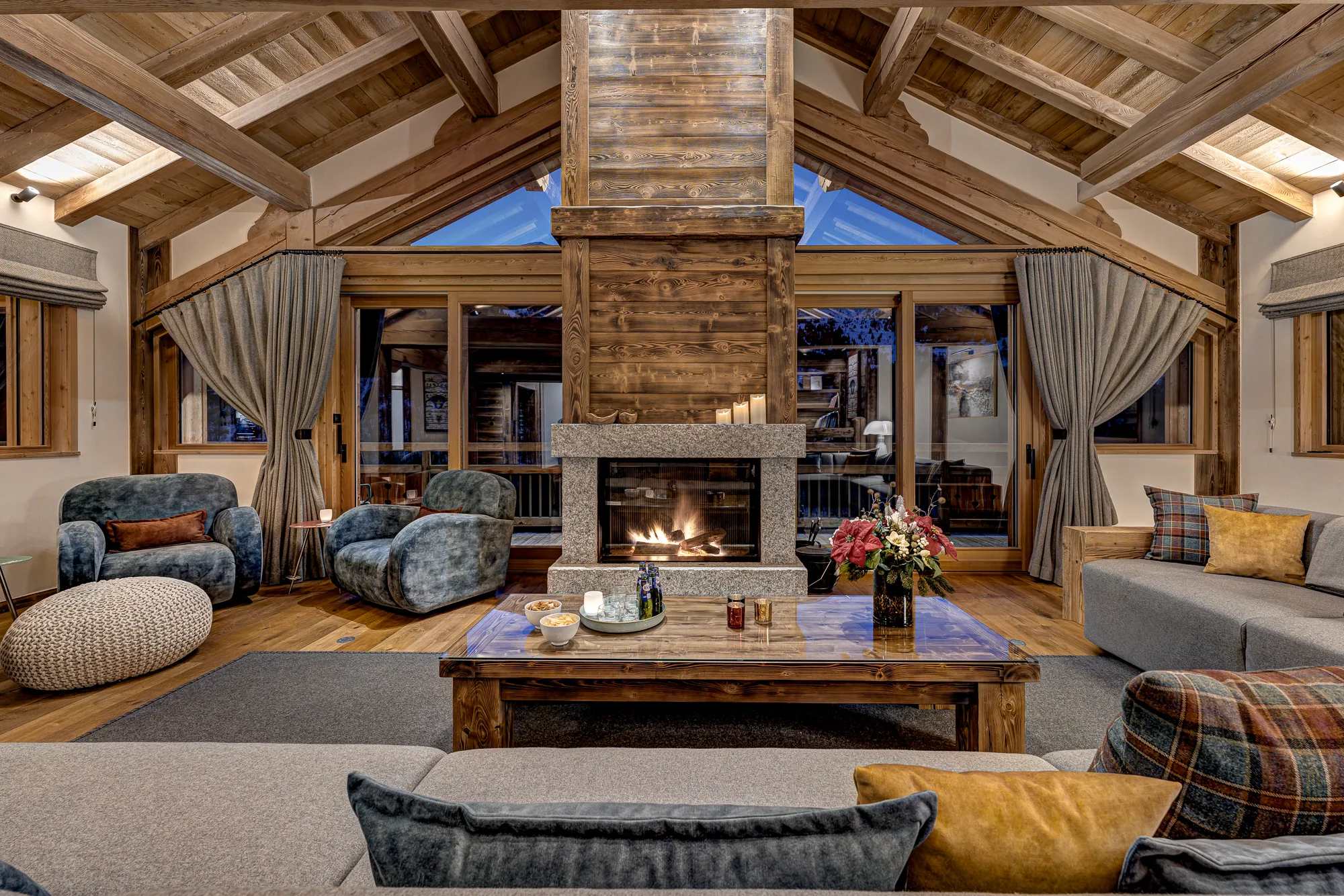 Cosy living room area at Chalet Myriam featuring comfy sofas with plaid cushions, a large lit stone fireplace and coffee table set under high vaulted ceilings with a glass wall behind
