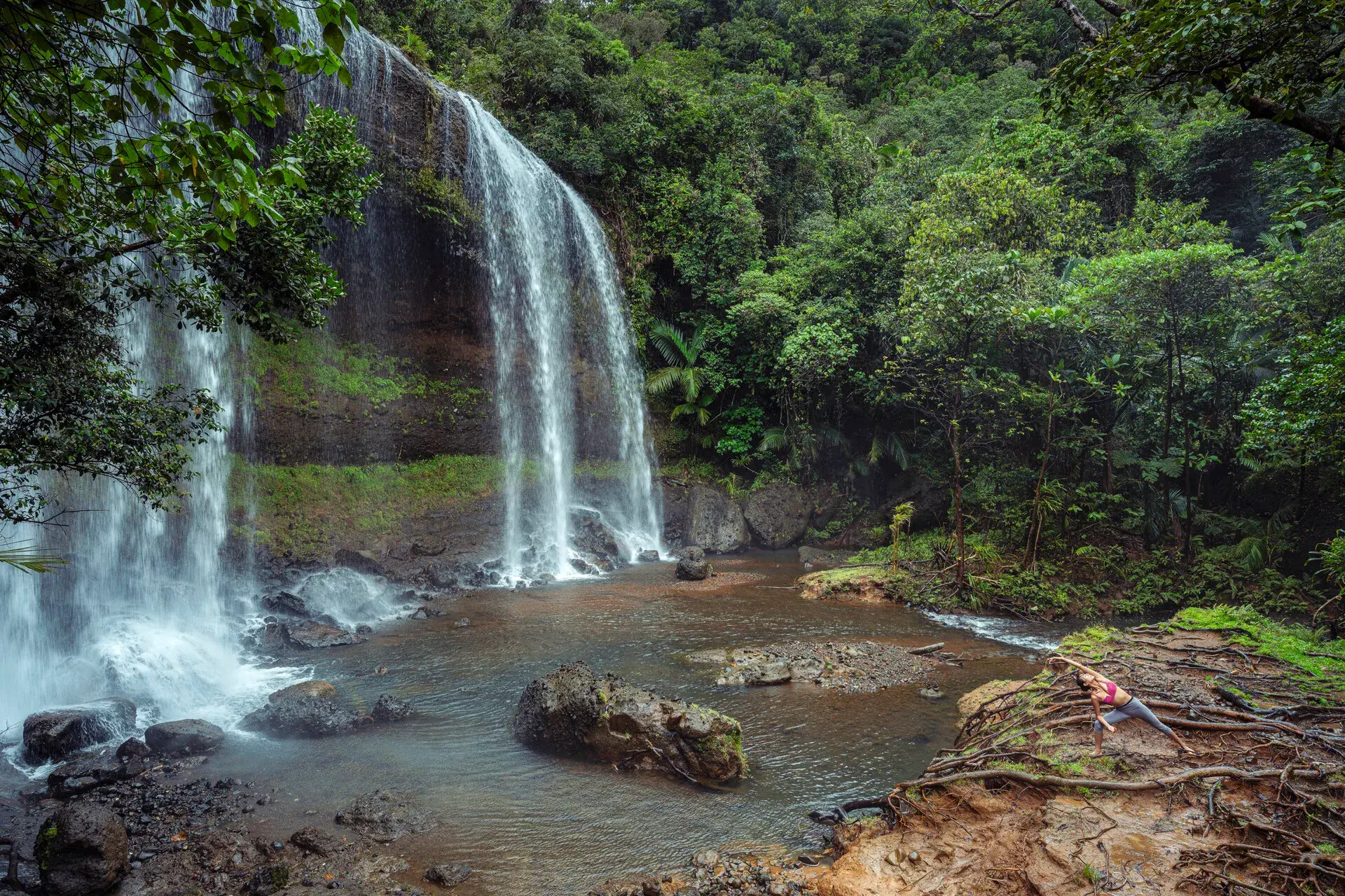 A large waterfall with streams gushing into a pool below surrounded by lush jungle with a person practicing yoga below