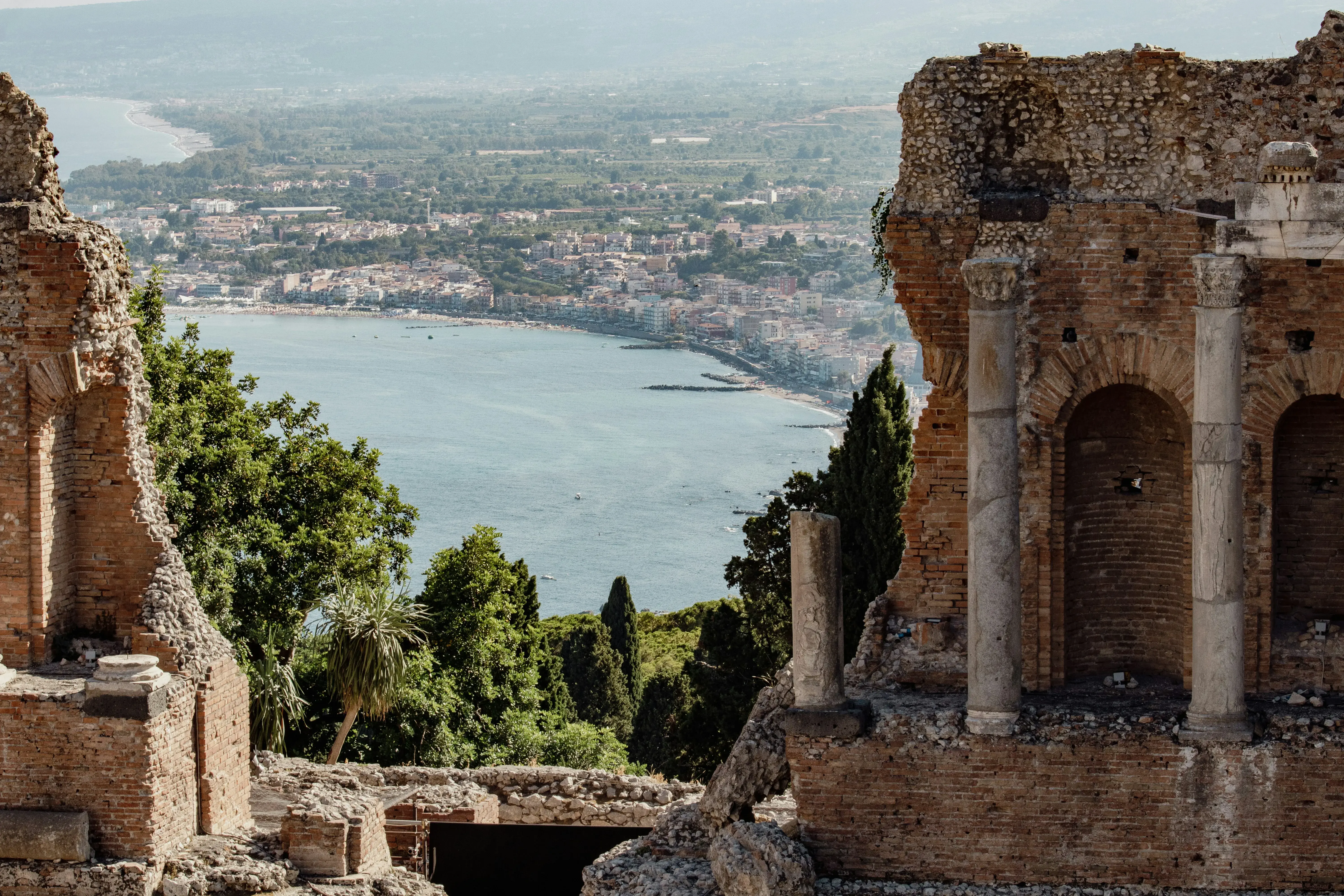 A view of the city of Taormina and the bay through the ruins of the ancient theatre of Taormina above green trees