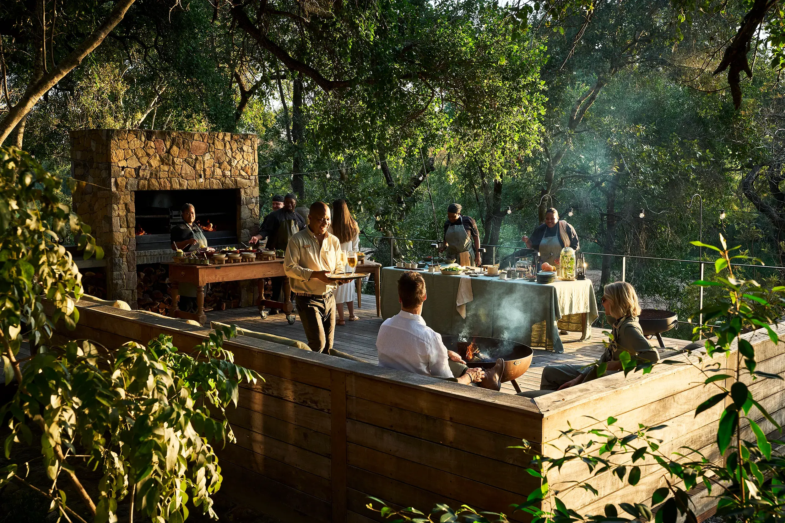Two guests attending an outdoor barbecue at Monwana Game Reserve featuring staff cooking behind well-stocked tables and an open fireplace with a waiter bringing the couple drinks