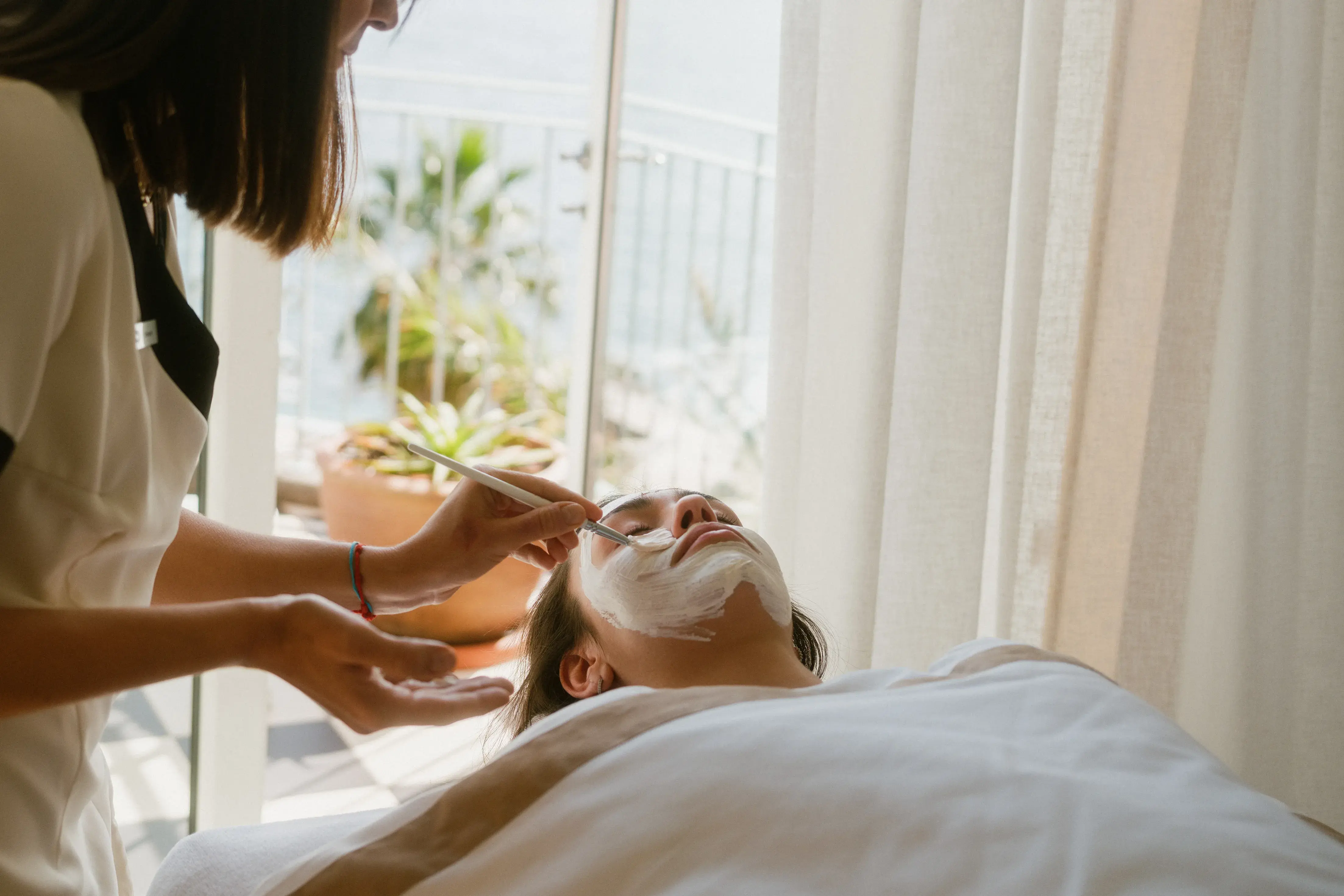 Person receiving a relaxing facial treatment with a brush mask application in a bright, serene spa setting.