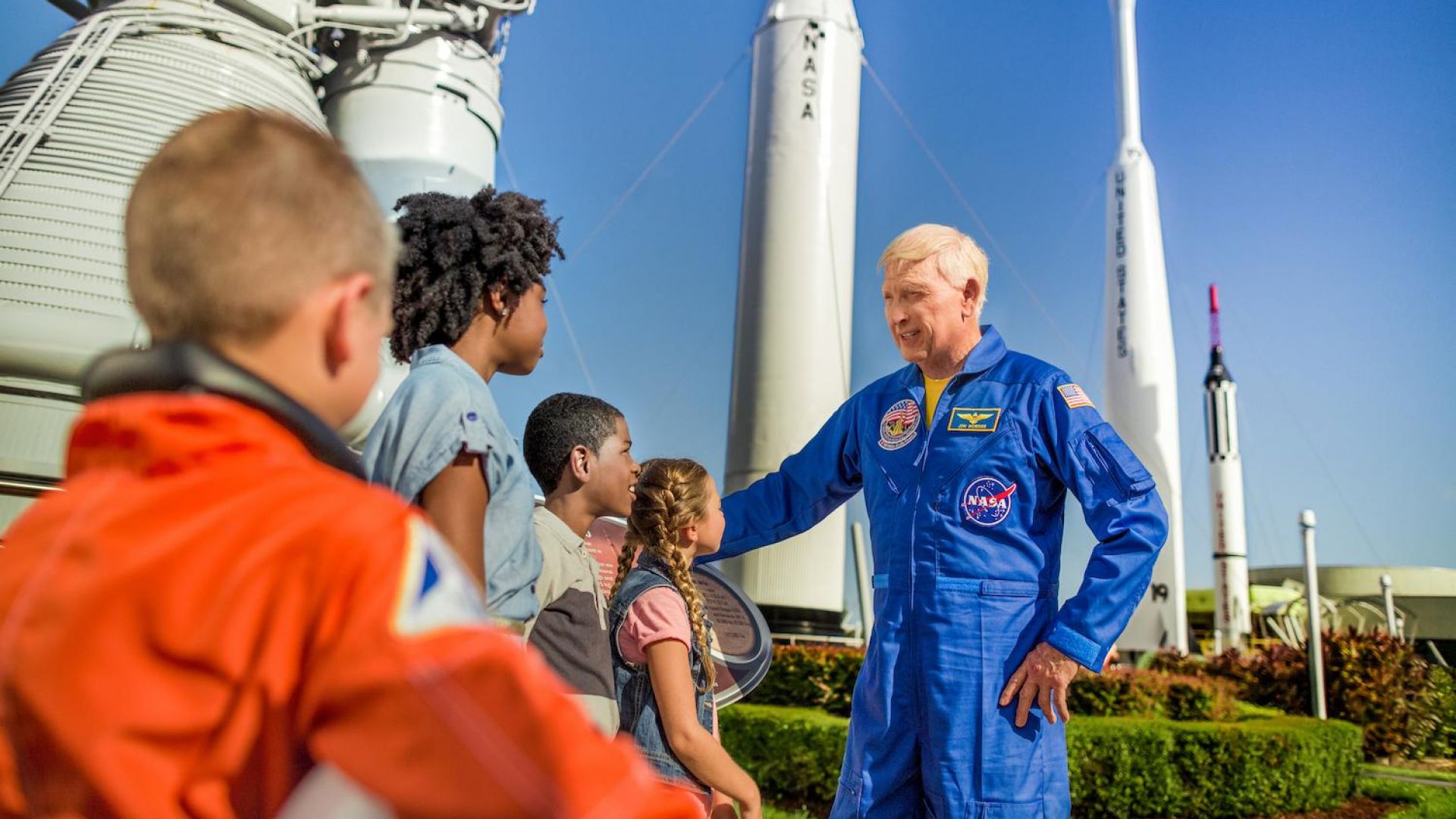 An astronaut in a blue NASA spacesuit speaks to children in front of rocket displays at Kennedy Space Center