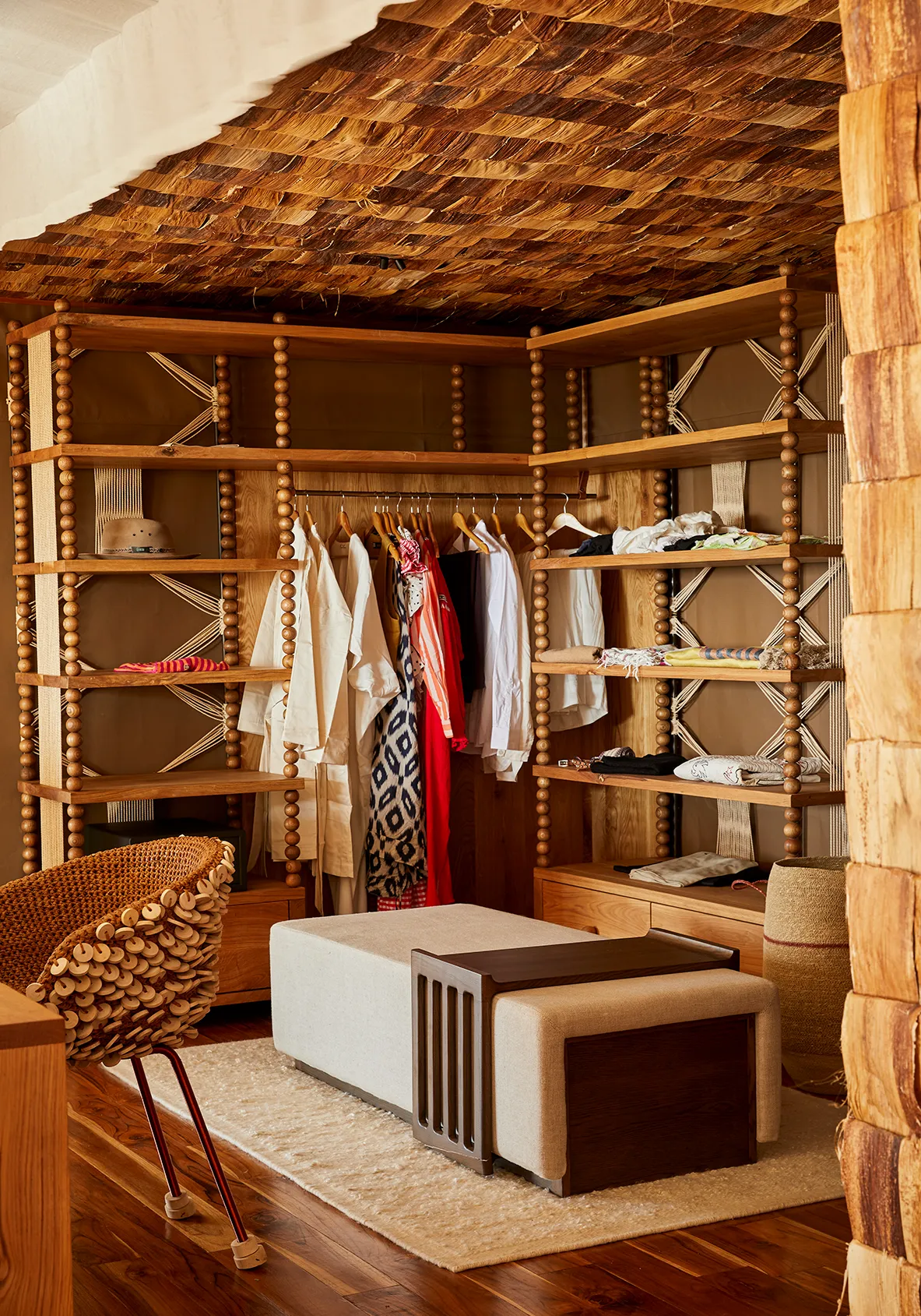 Open wooden walk-in wardrobe with shelving, hanging clothes, woven accents, and a cushioned bench.