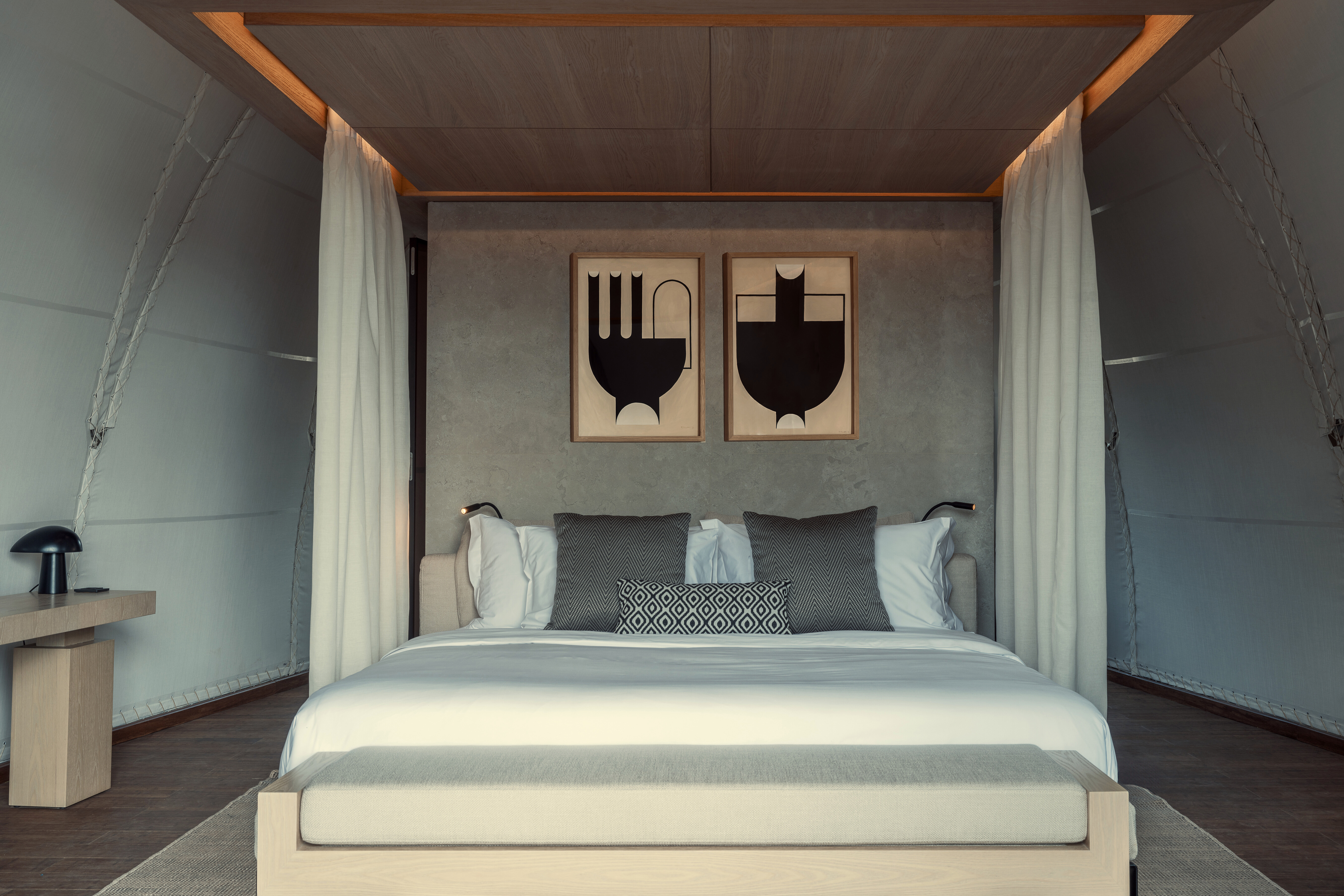 Large plush bed in shades of white and grey with sheer white curtains and two geometric prints behind