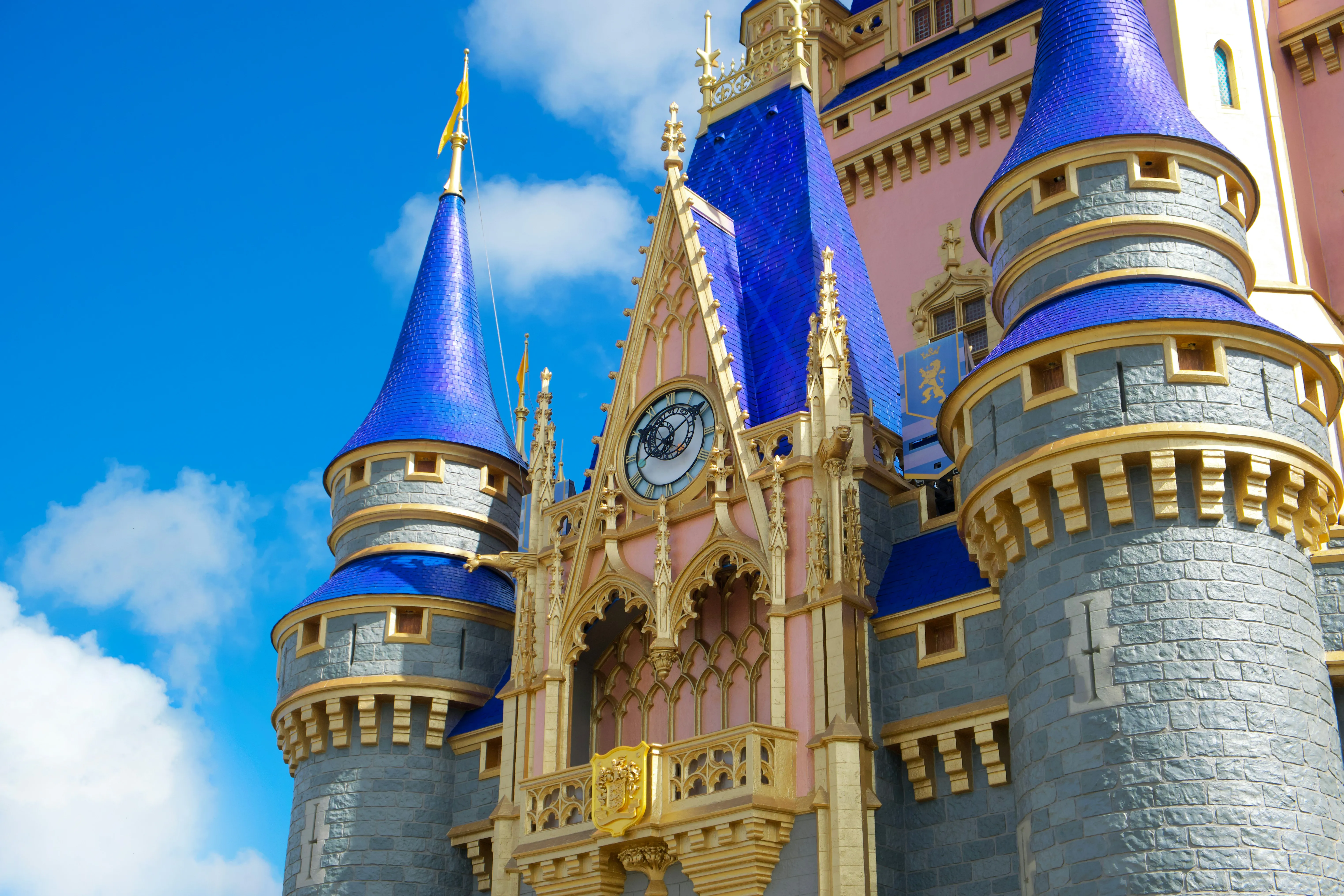 Close-up view of a fairytale-style castle with blue spires, gold detailing, and a central clock tower against a bright blue sky.