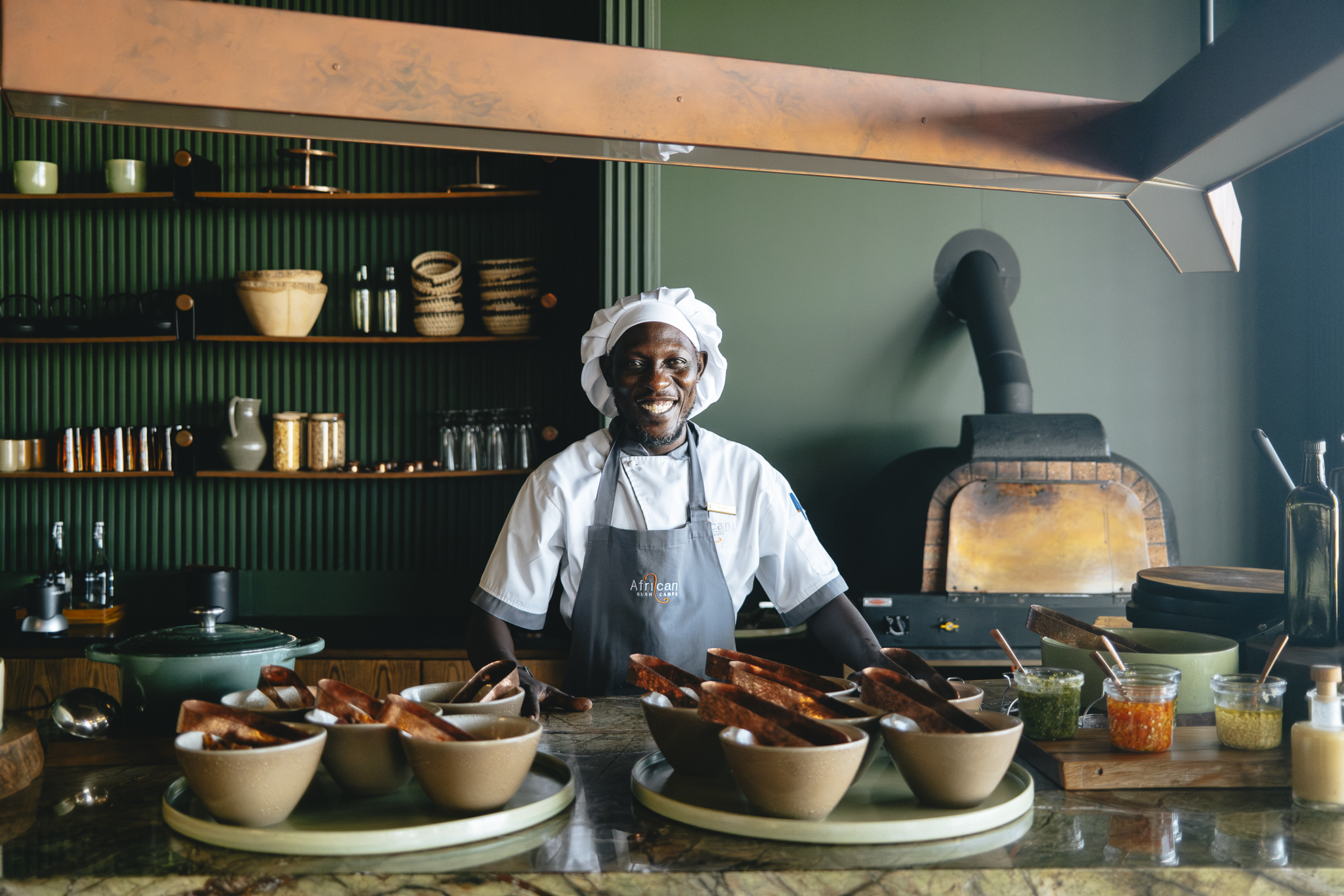 The chef at Lolebezi standing smiling in his kitchen surrounded by pots and pans with a woodfired oven in the background