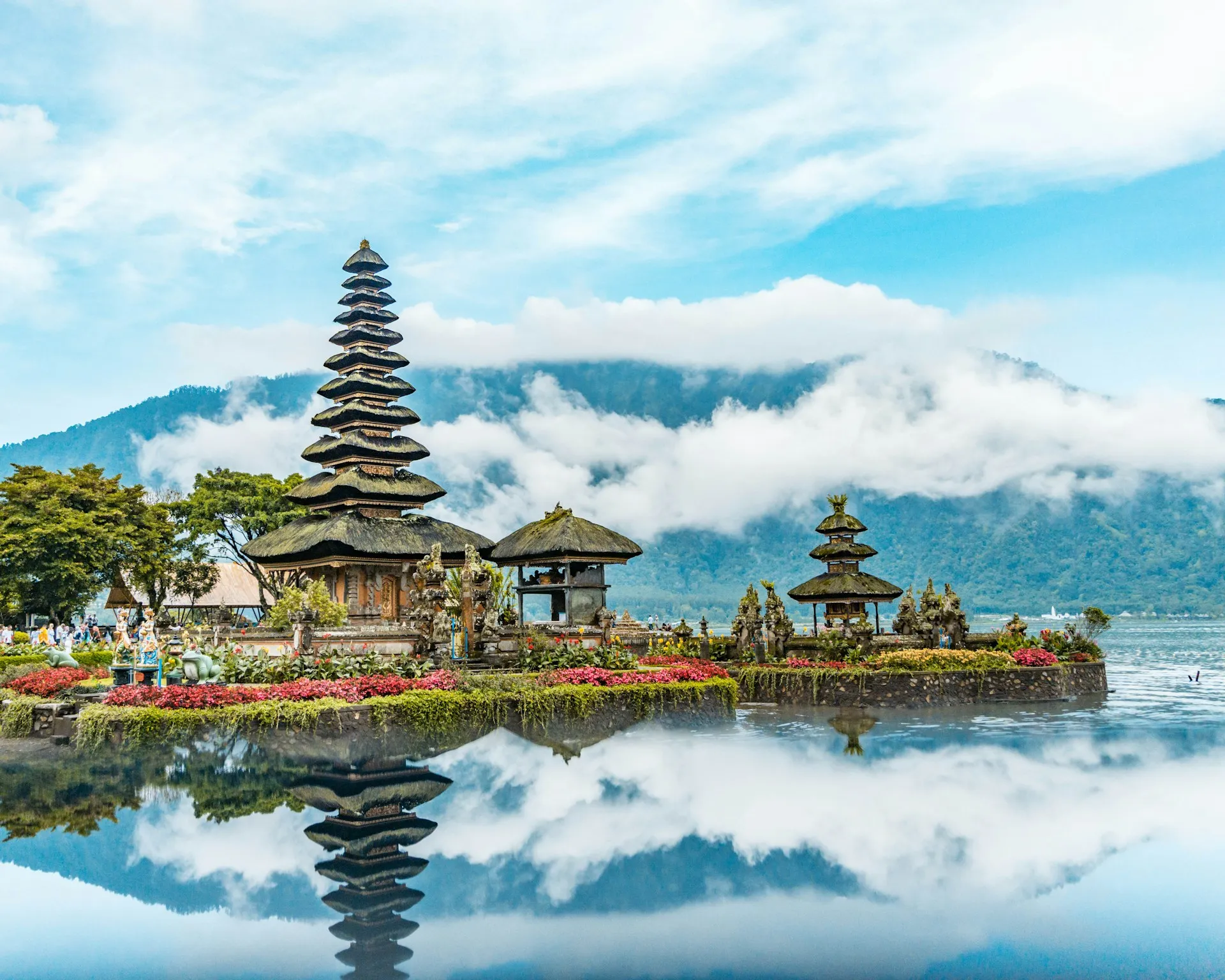 A traditional Balinese temple with tiered roofs is reflected in a calm lake, surrounded by greenery and mountains under a blue sky.