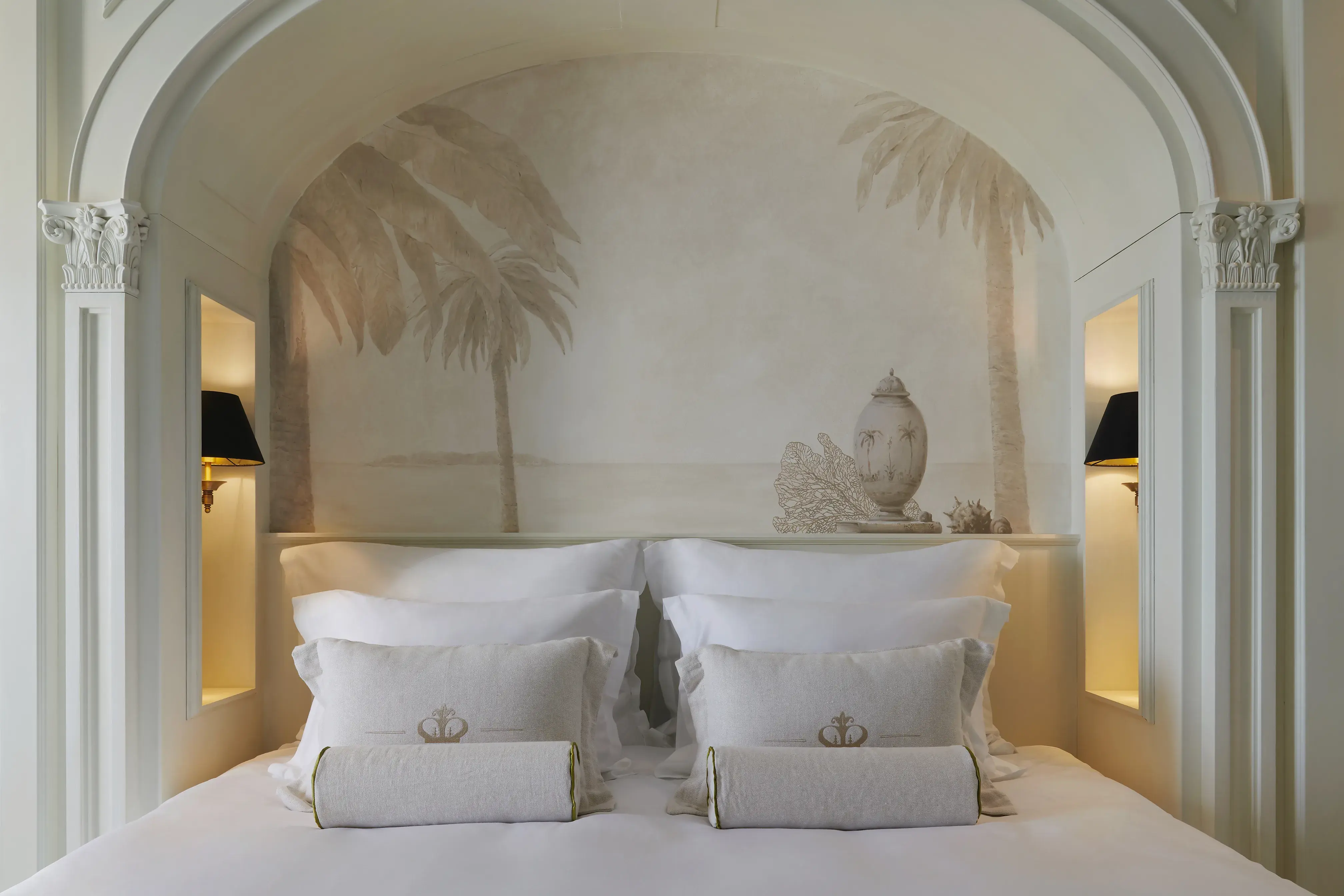 A plush bed at Palace Elisabeth in Croatia with white linens set beneath a decorative arch with a pale mural of palm trees behind