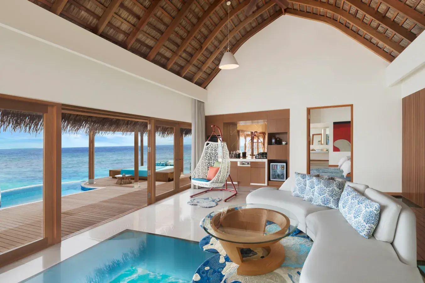 The living room of an Overwater Two Bedroom Villa at W Maldives featuring soaring vaulted ceilings, floor-to-ceiling glass doors, a private oceanfront deck with infinity pool, and panoramic views of the turquoise Indian Ocean