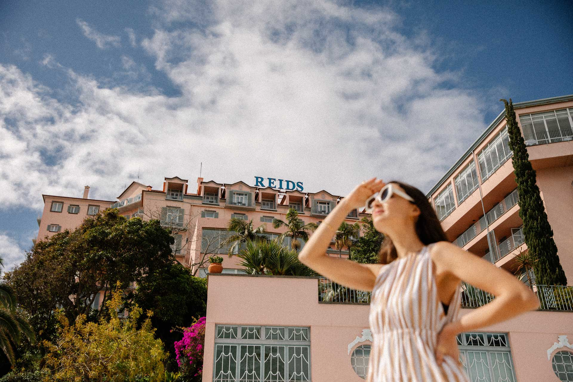 Exterior view of Reid’s Palace hotel with a guest in the foreground and lush gardens under a bright sky.