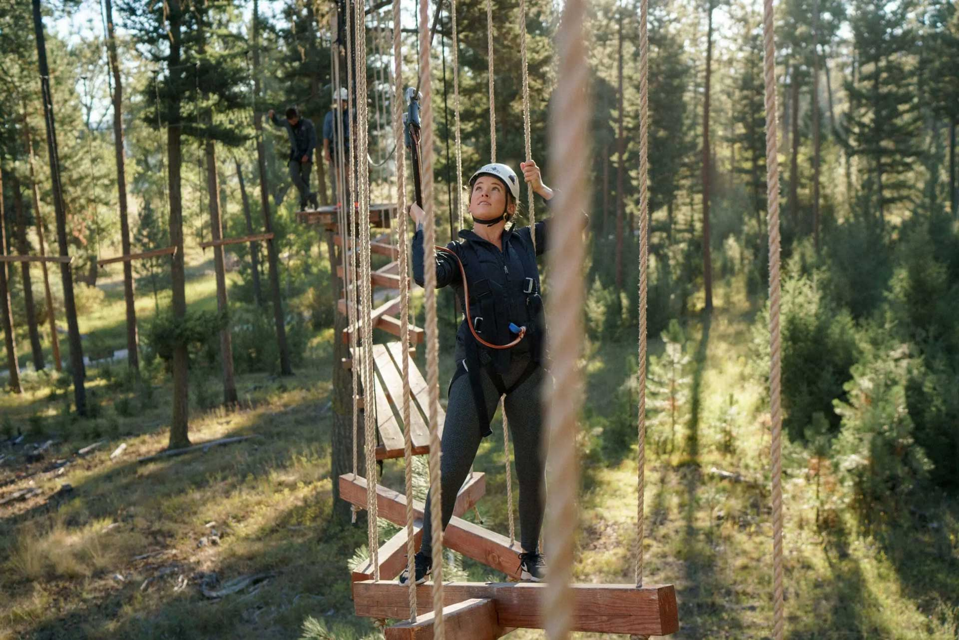 Person navigating a high ropes course between trees while secured with a harness in a forest setting.