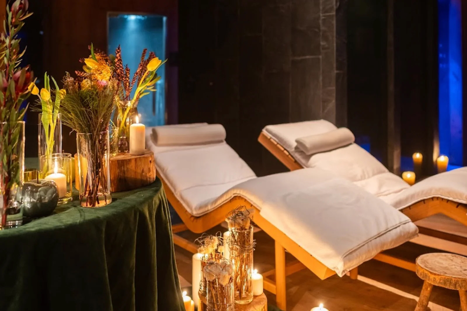 Loungers in the spa beside a table with green cloth and lit candles and vases of dried flowers on top
