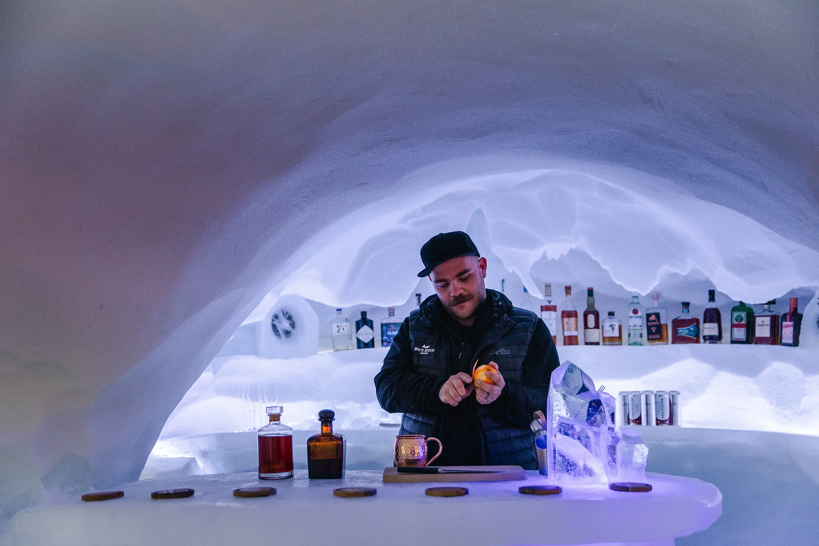 Bartender preparing a drink inside an atmospheric ice bar with bottles displayed along carved snow shelves.