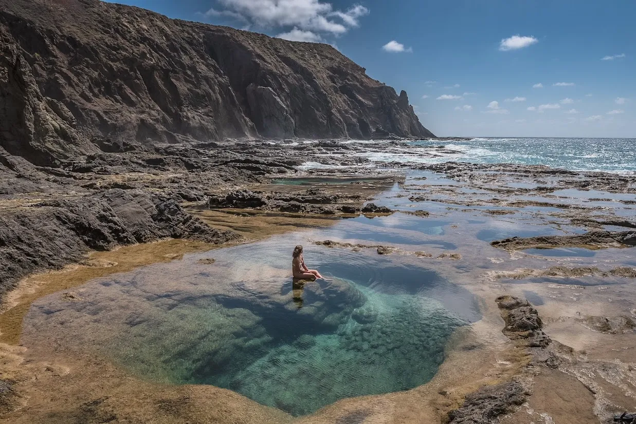 Person sitting in a natural rock pool beside a rugged coastline and ocean cliffs.