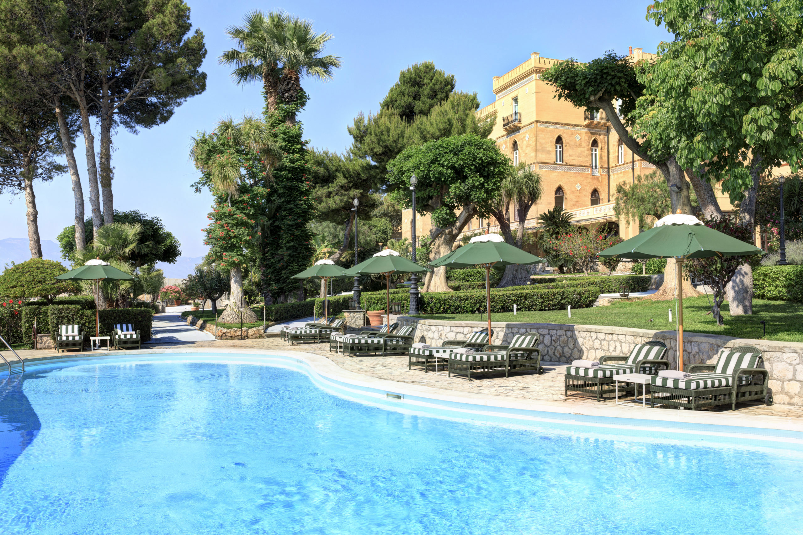 A luxurious outdoor pool area with striped sun loungers and green umbrellas, surrounded by lush trees and a stately yellow building in the background.