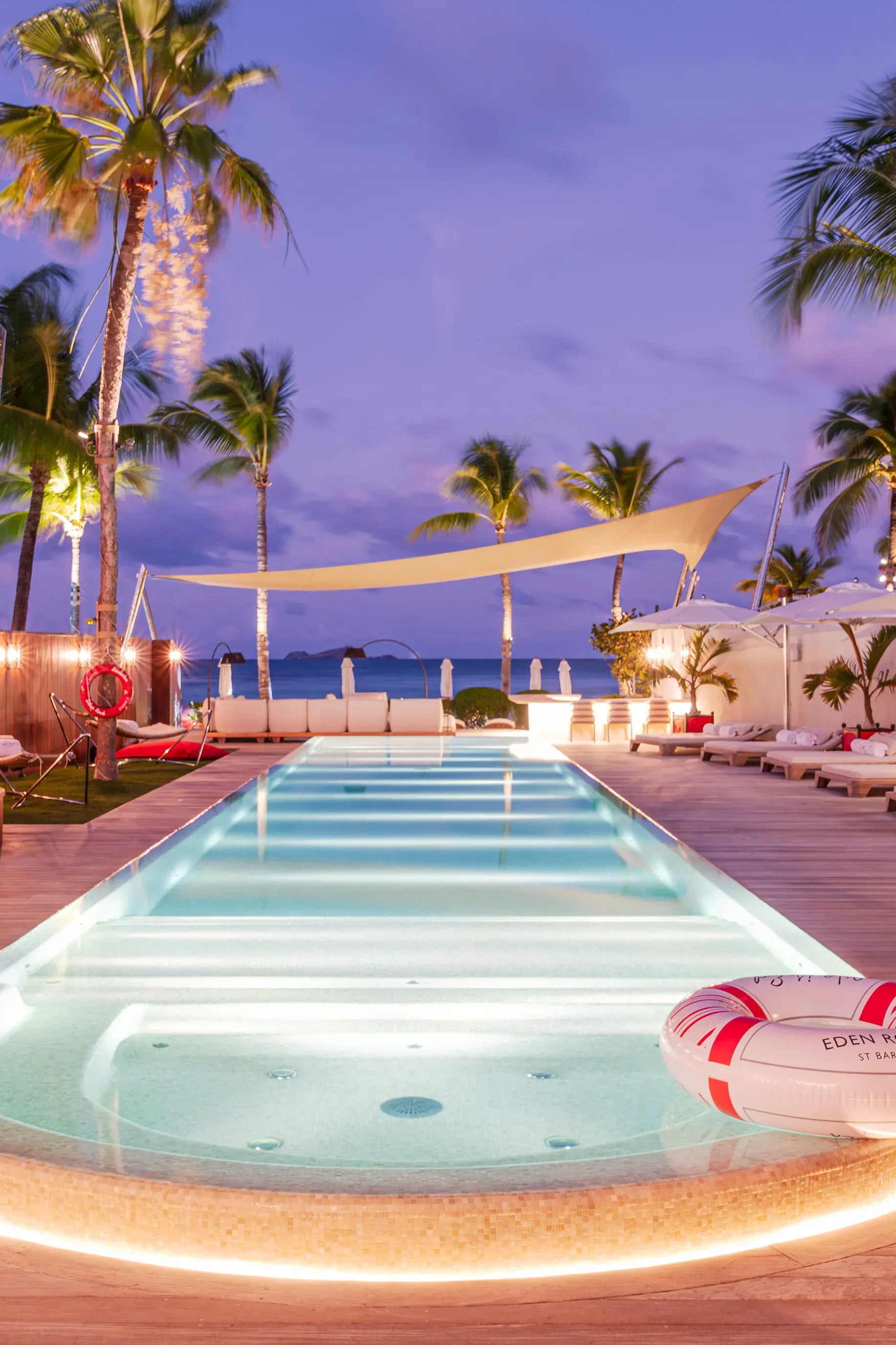The long swimming pool of Villa Rockstar at Eden Rock in St Barths lit up at night, featuring a branded rubber ring and white sun loungers looking out to sea