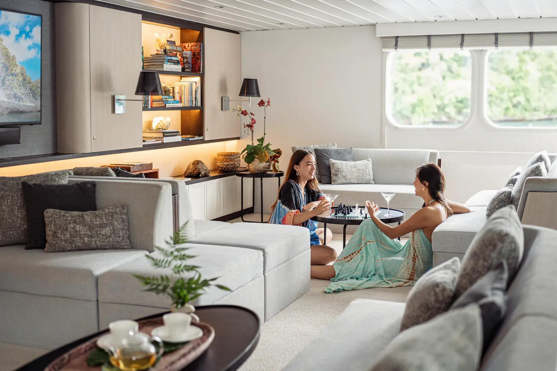 Two guests of Four Seasons Explorer Palau drinking cocktails and playing chess in a chic lounge area with pale sofas, a large television and ornaments and books on shelves