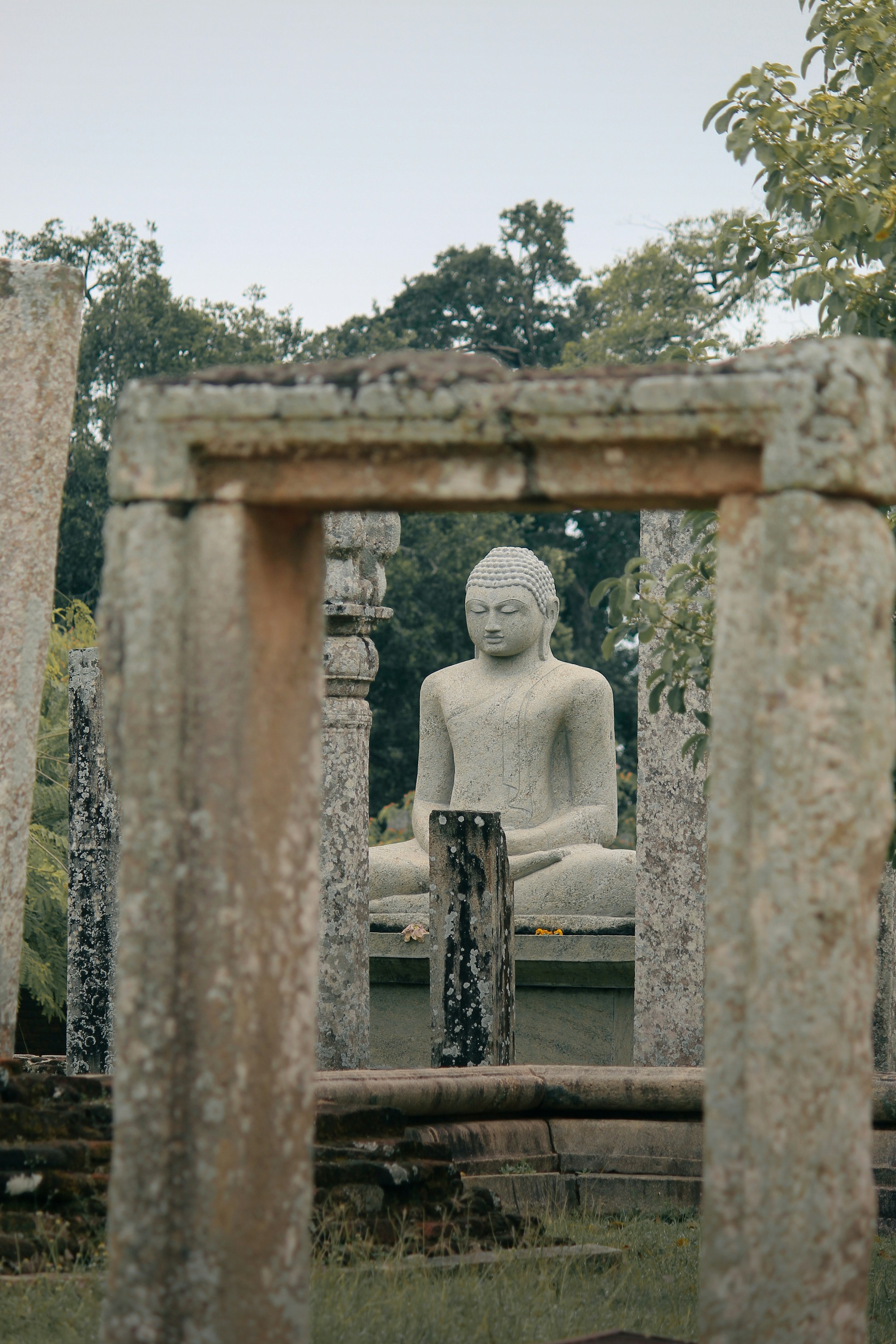 Ancient stone Buddha statue framed by weathered pillars in a historic archaeological site surrounded by lush greenery.