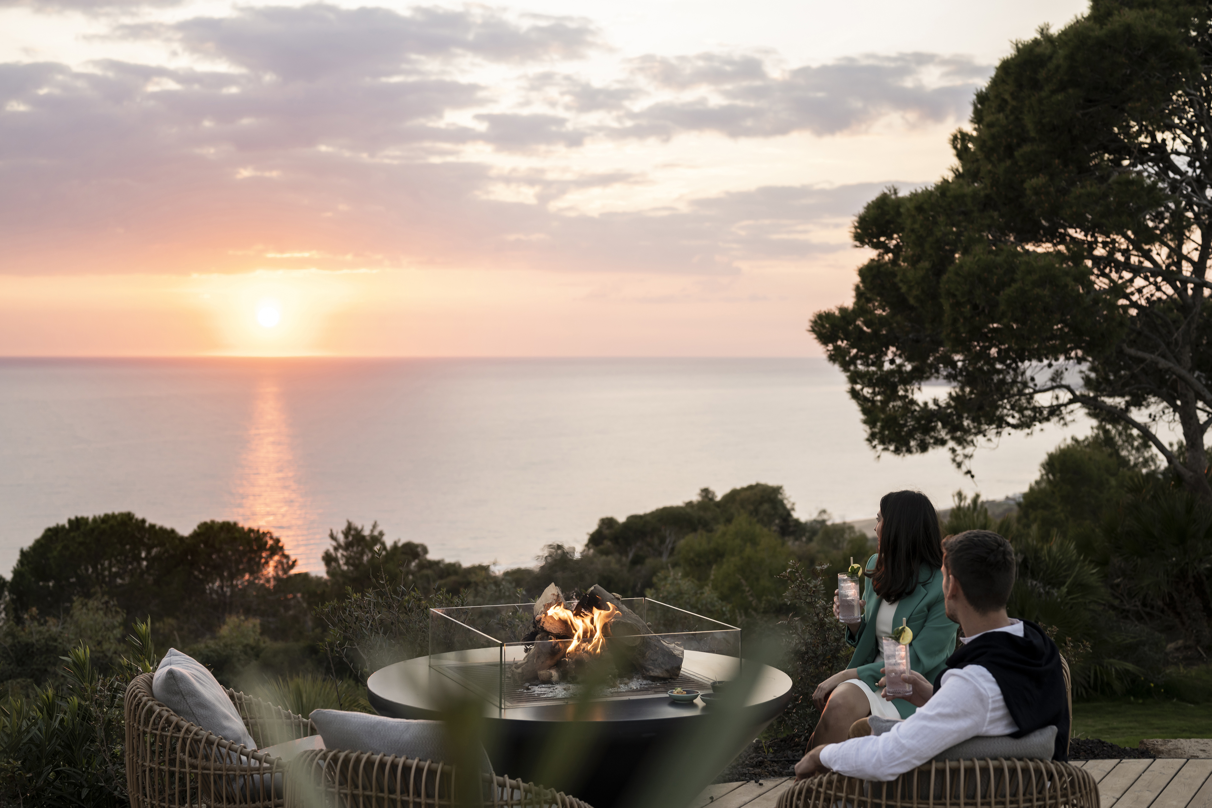 Couple with drinks seated around a lit outdoor fireplace looking out to sea and the sunset on the horizon