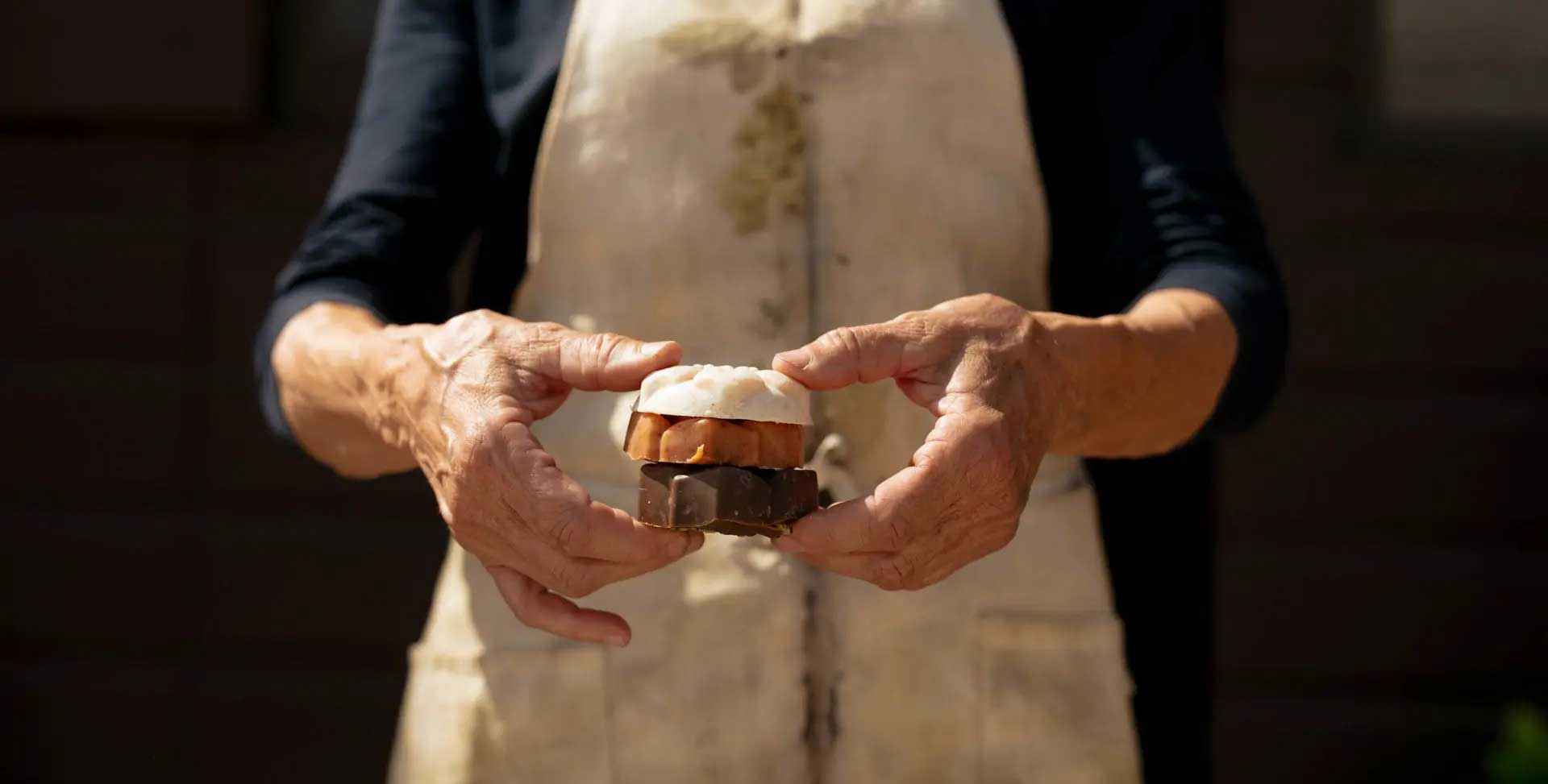 Close‑up of hands holding a stack of artisanal soaps in natural tones against a rustic wooden backdrop.