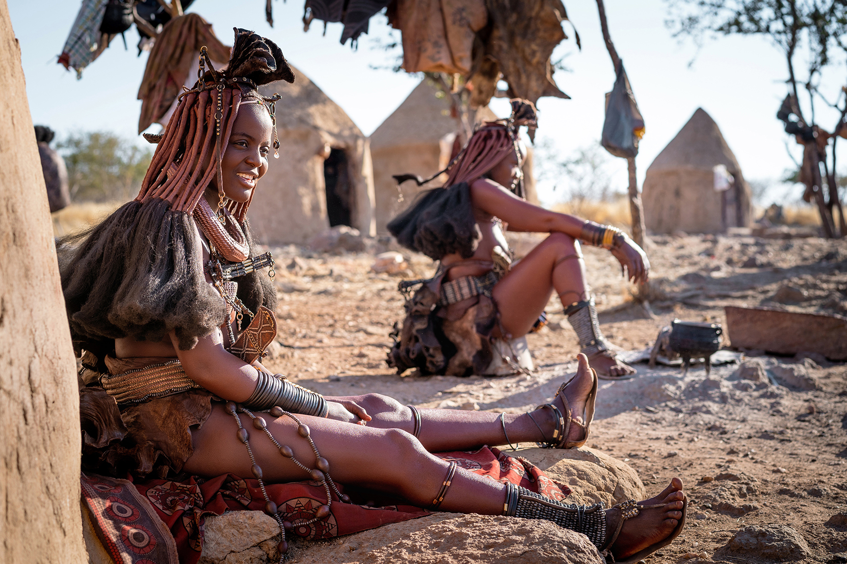 Two Himba women sat smiling in traditional dress in their village