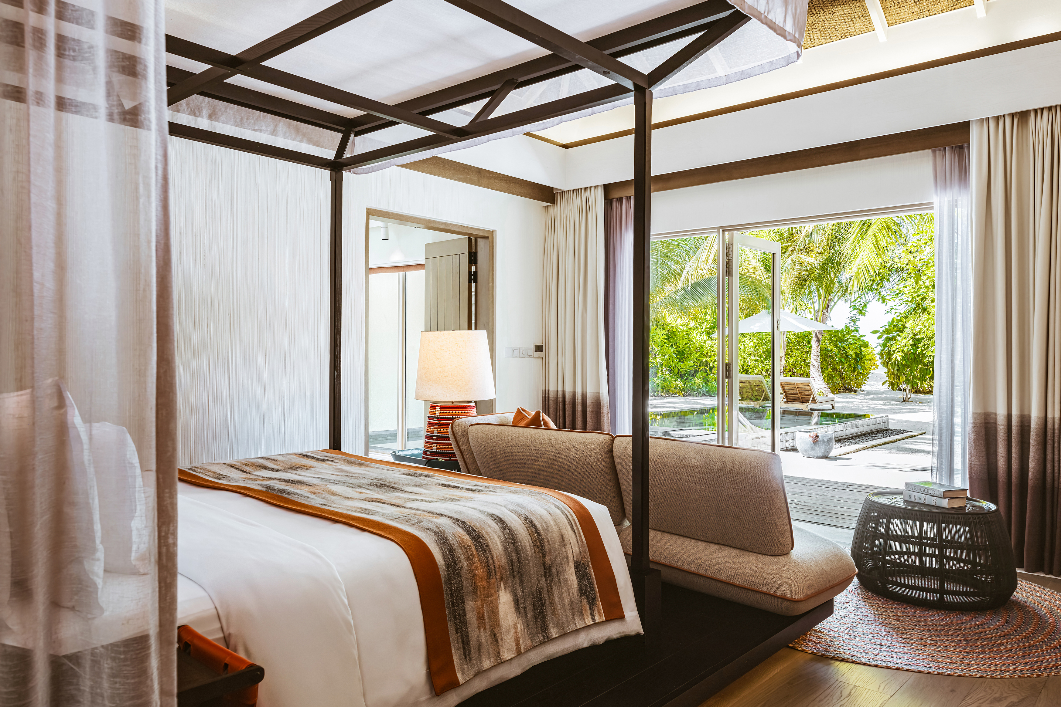 Luxurious InterContinental Maldives villa bedroom with canopy bed, modern decor, and view of private pool and lush garden on the beach through sliding glass doors.