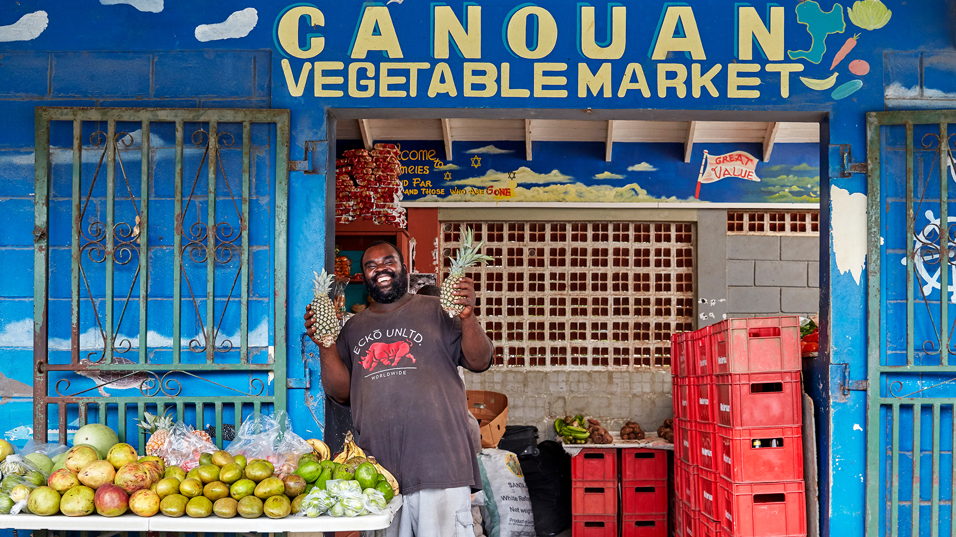 A man posing with pineapples in front of a blue market building with the words Canouan vegetable market written on it