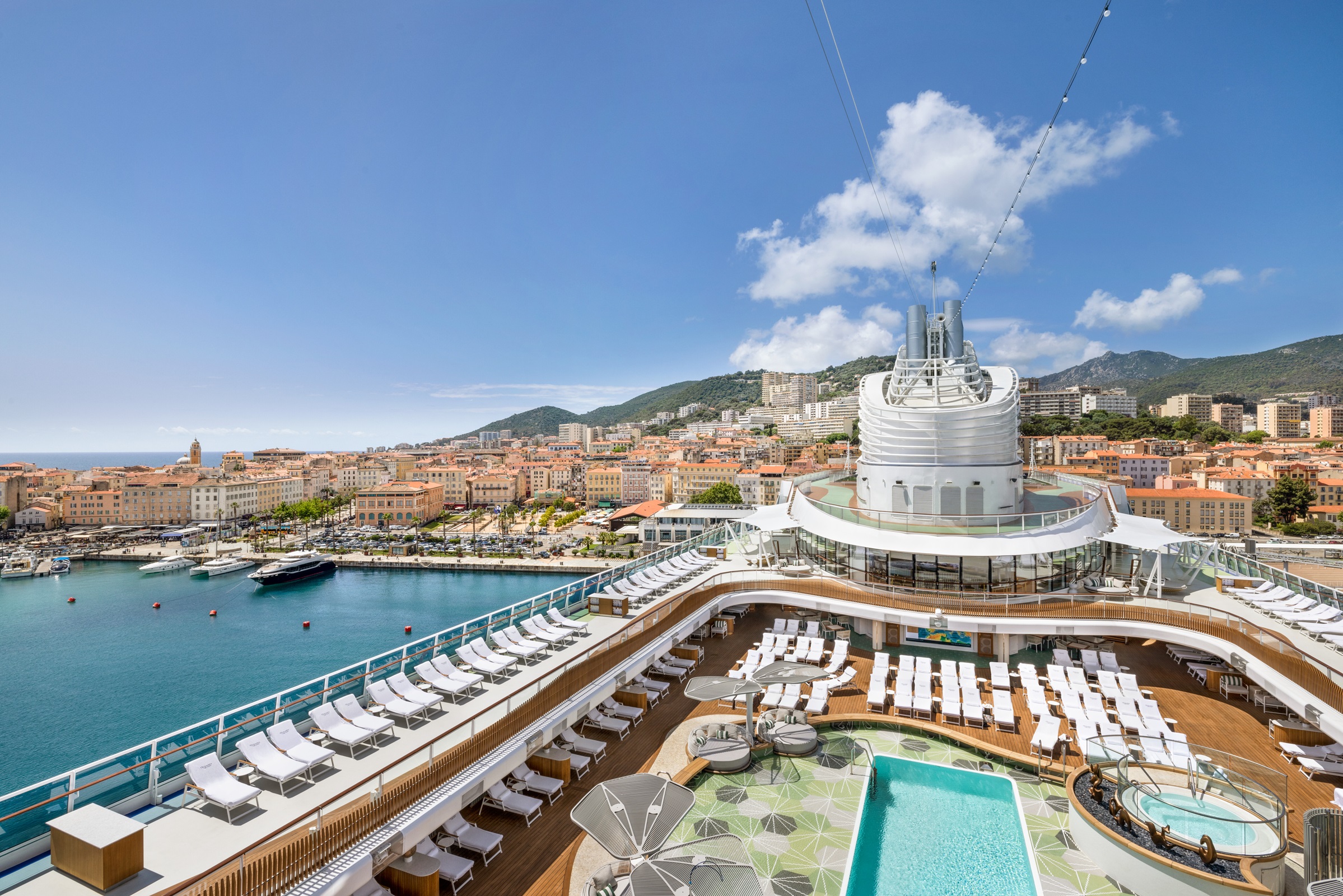 Oceania Cruises ship docked at port with pool and lounge chairs on deck, city and mountains in the background.